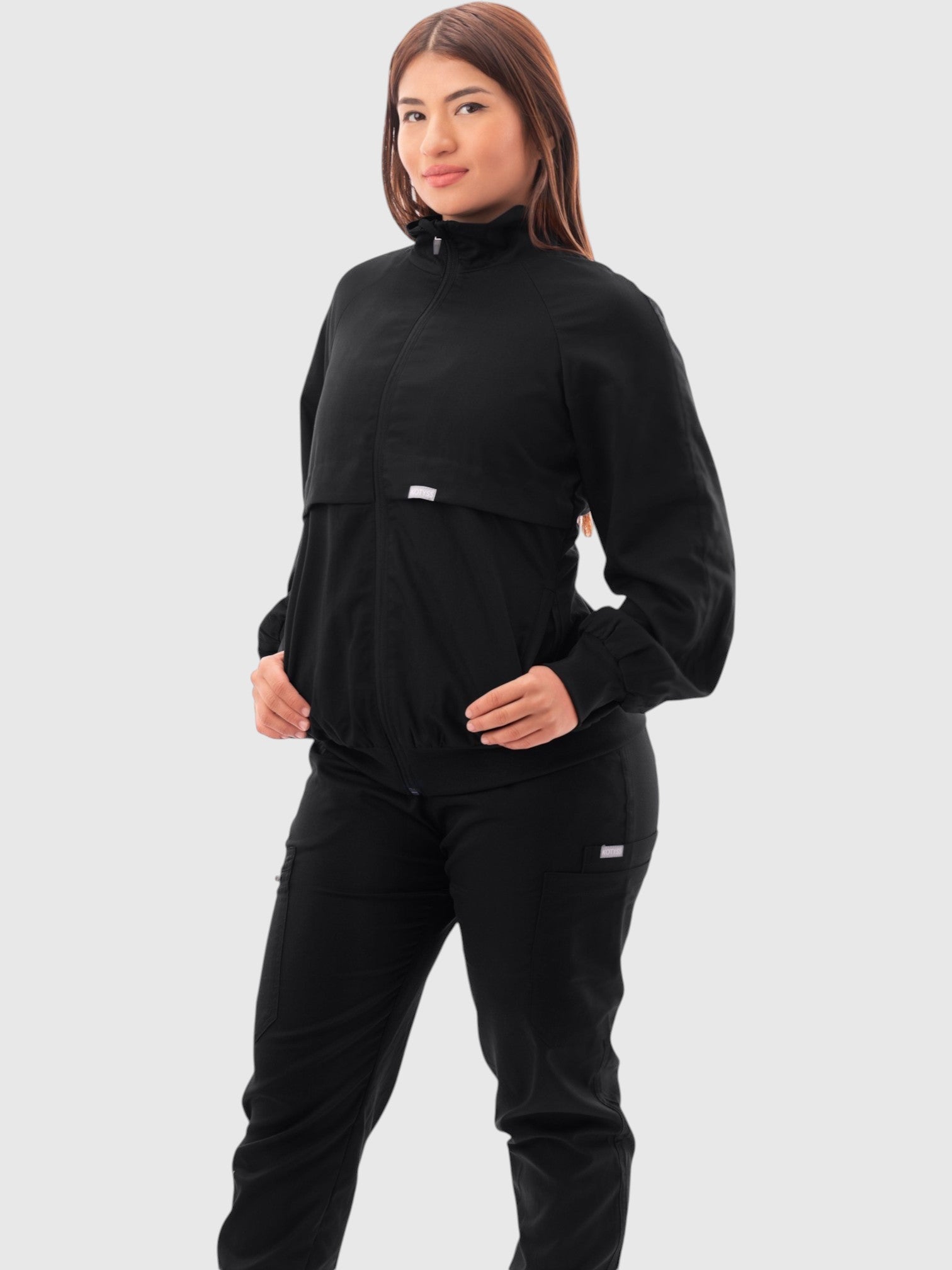 A woman wearing a black Kotyss scrub jacket with a high windbreaker neck, ribbed cuffs, and functional pockets.