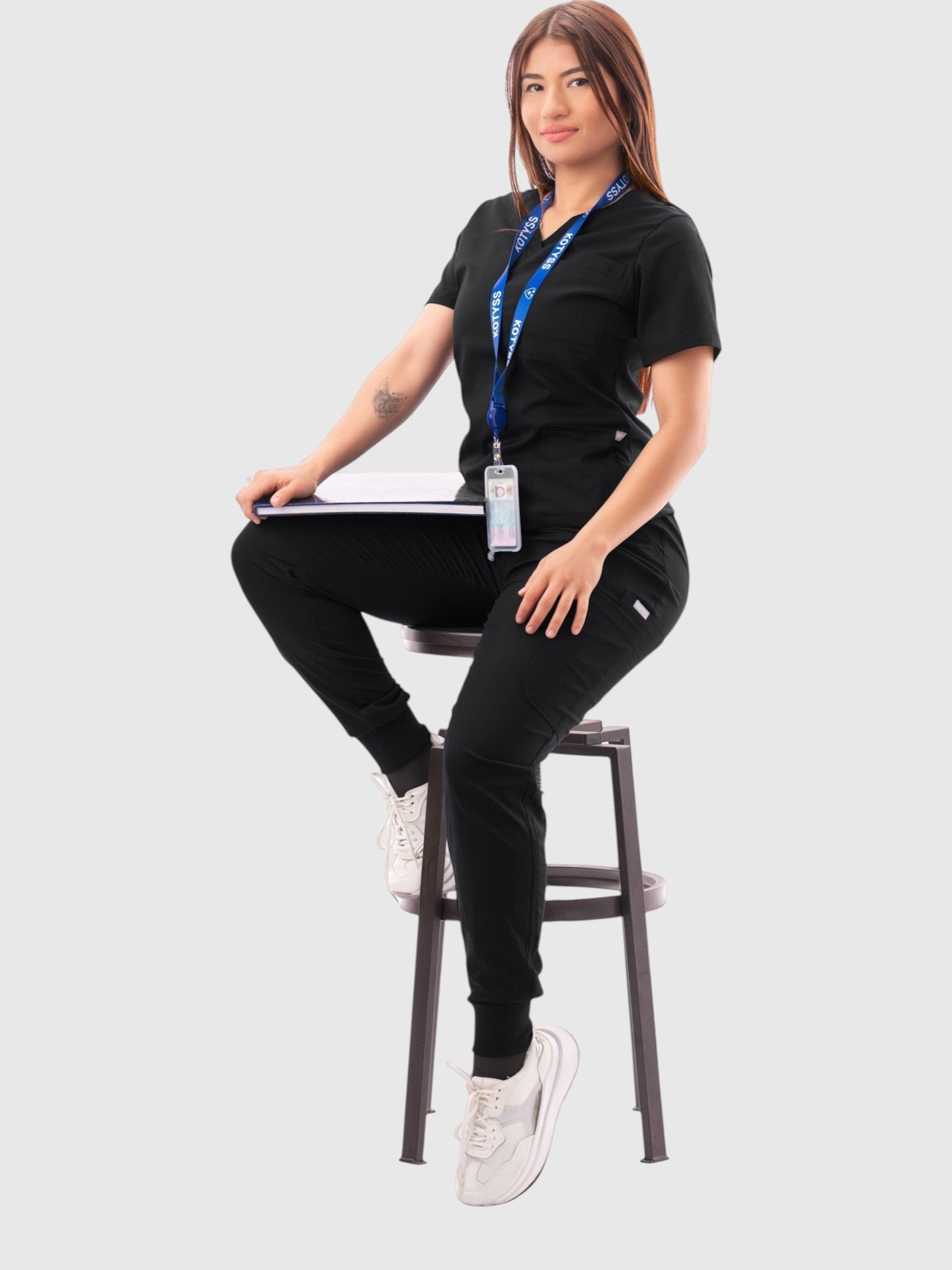 A woman wearing a black Kotyss V-neck scrub top and multi-pocket jogger pants, sitting on a stool in a studio.