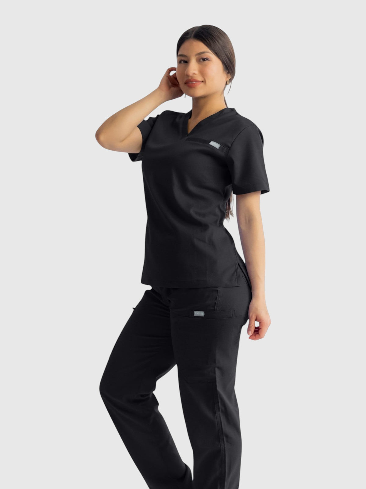 A woman wearing Kotyss black straight cut medical scrub pants and matching v-neck top, featuring 8 pockets.