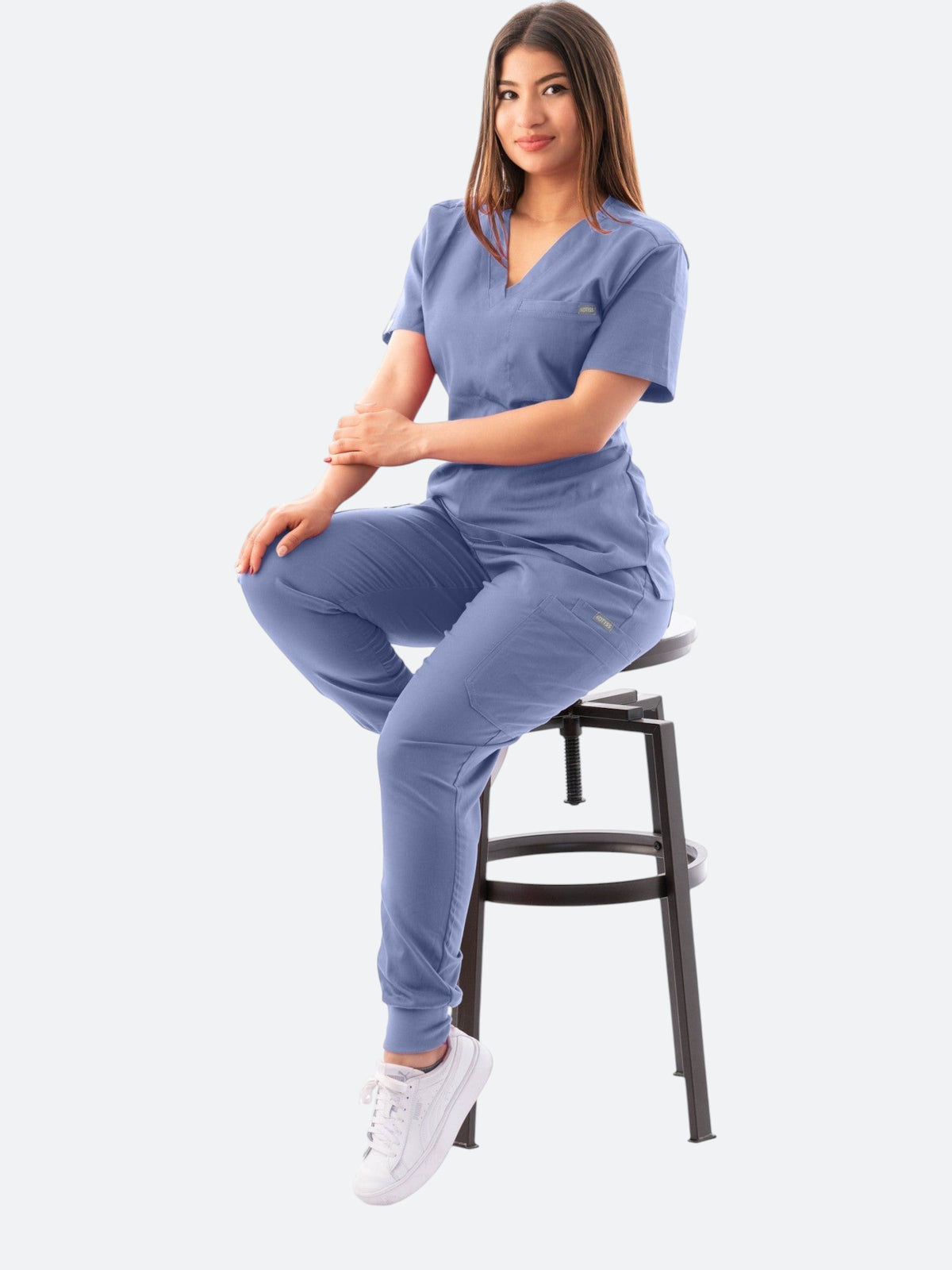 A woman wearing a blue Kotyss V-neck scrub top and jogger pants set while sitting on a stool.