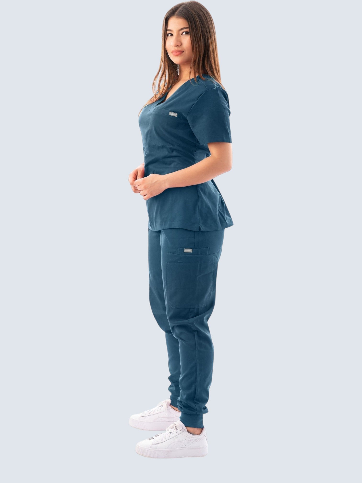 A woman wearing a Kotyss dark blue V-neck scrub top and matching jogger pants with multiple pockets.