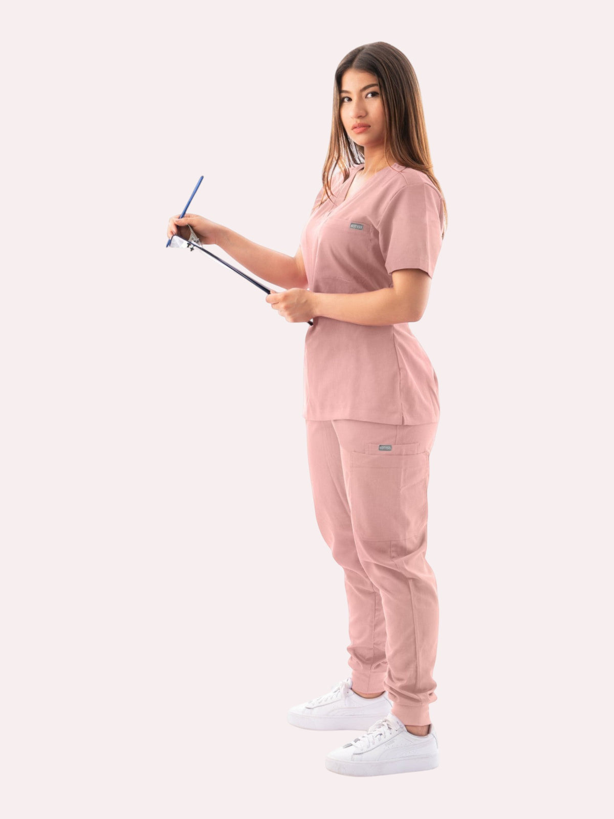 Kotyss women's blush pink V-neck scrub top and jogger pants set worn by a model holding a clipboard.