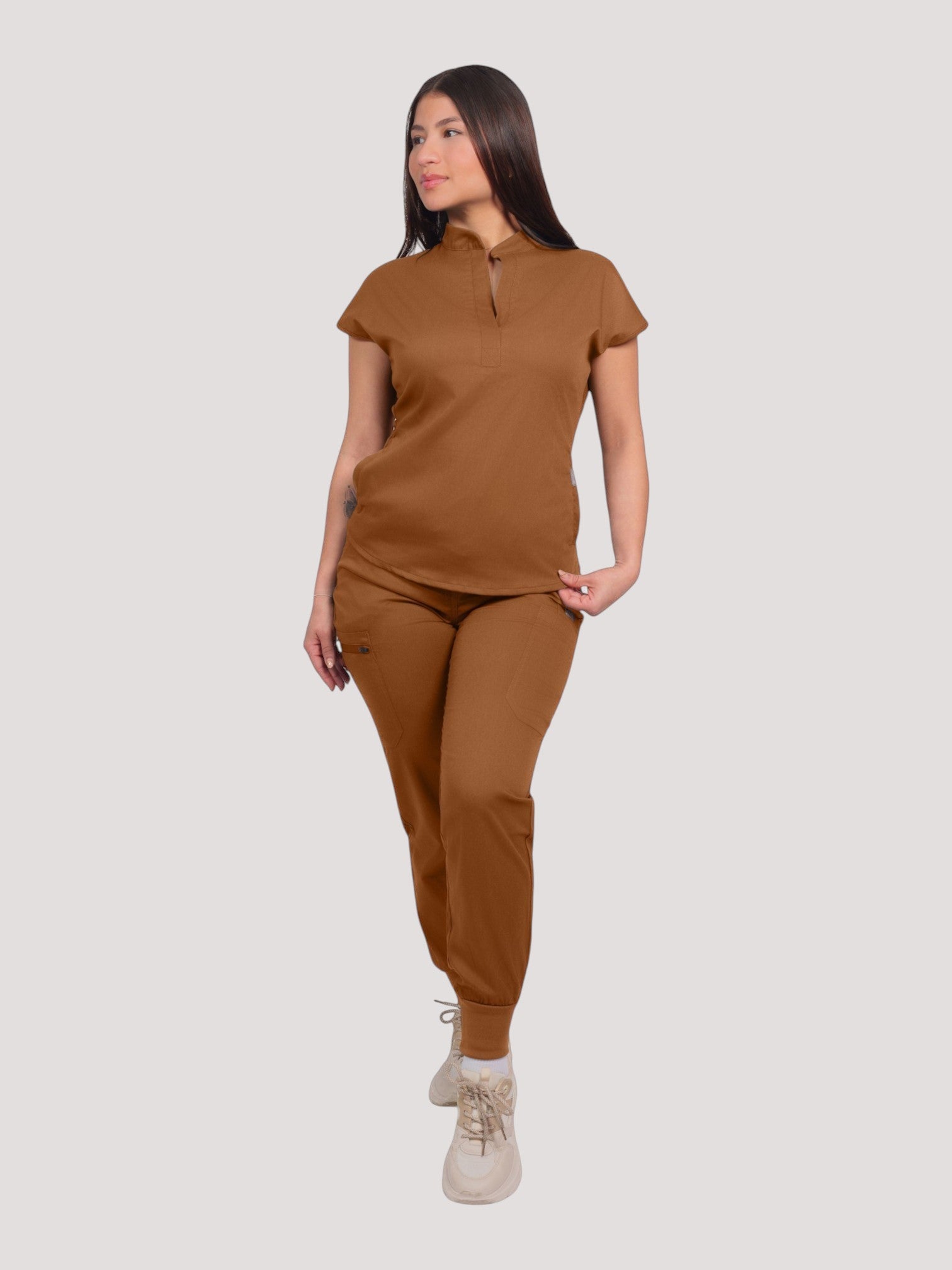 Model wearing a brown Kotyss collared scrub top and matching jogger pants with multiple pockets in a studio setting