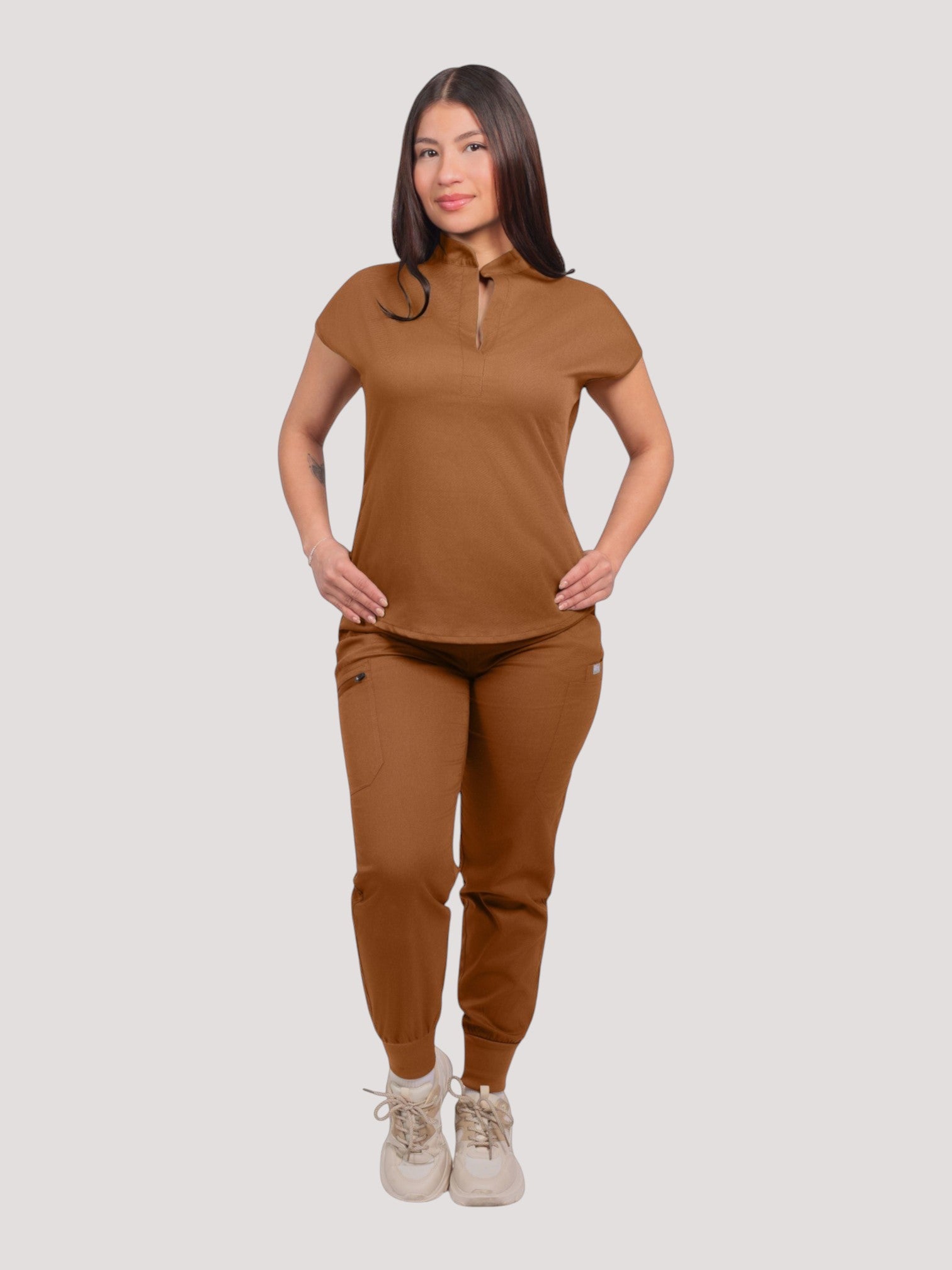A woman wearing a brown Kotyss collared scrub top and matching multi-pocket jogger pants in a studio setting.