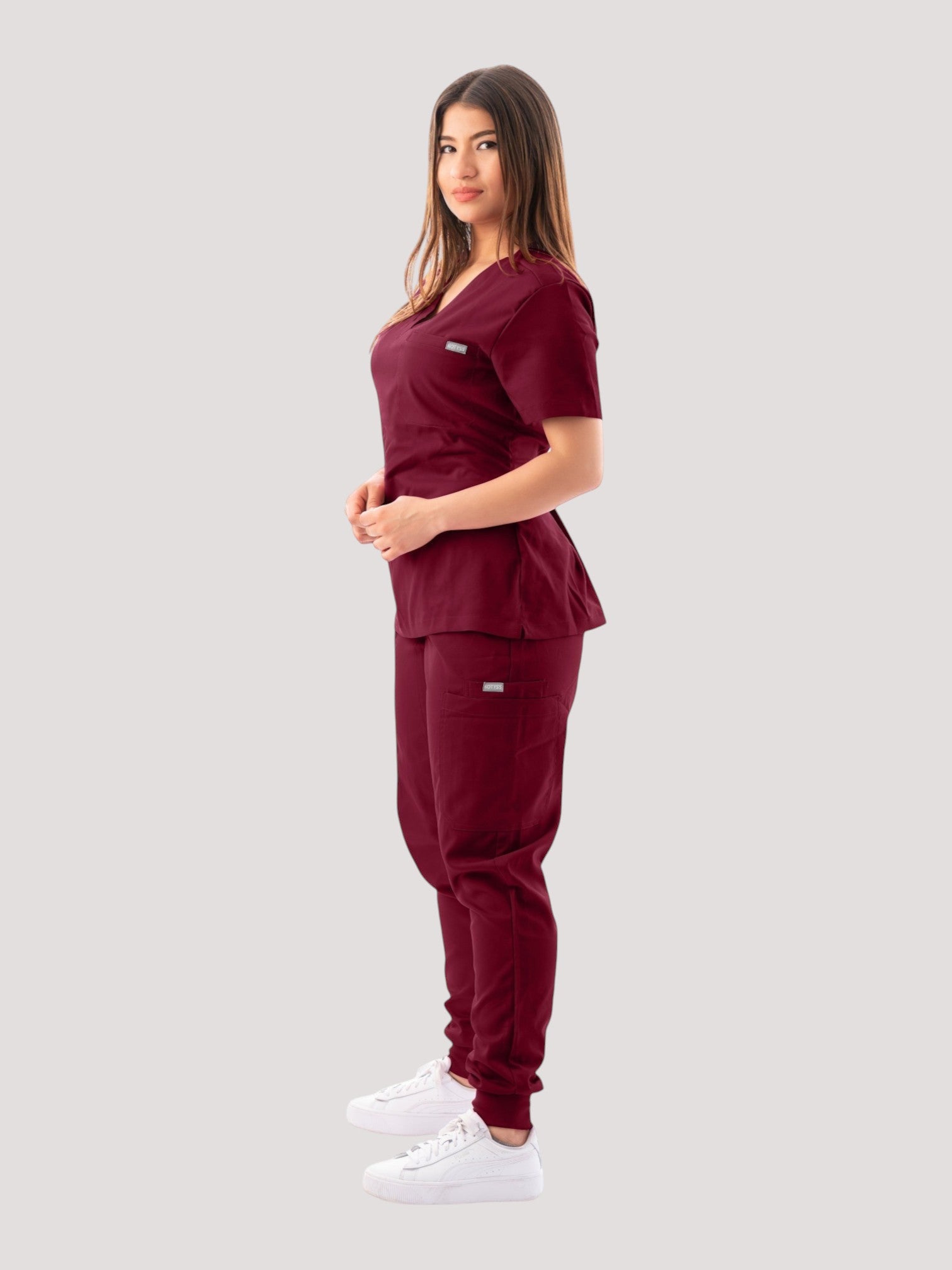 A woman wearing a Kotyss burgundy V-neck scrub top and matching jogger pants, side view against a white background.