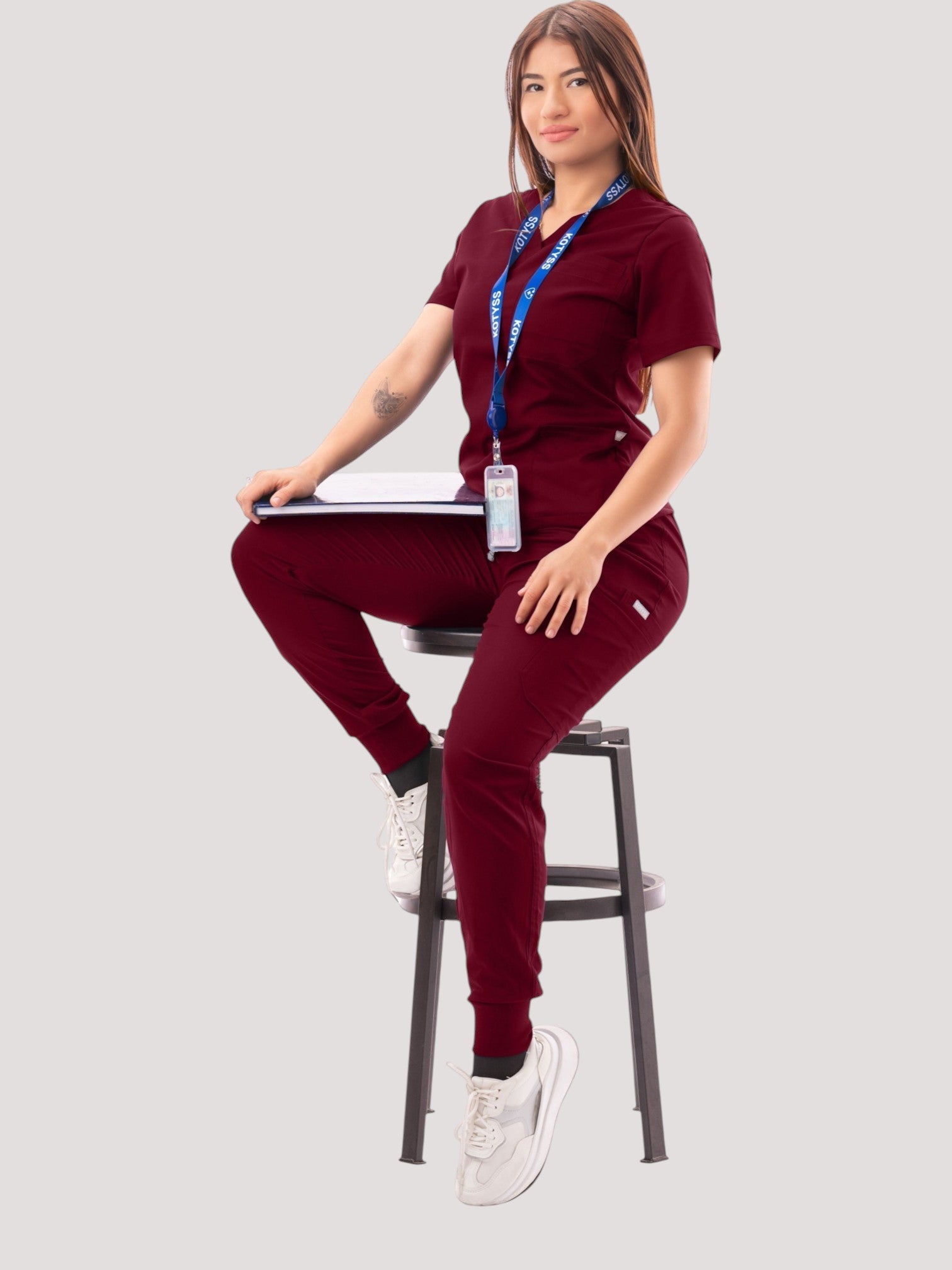 Woman wearing a burgundy Kotyss V-neck scrub top and jogger pants set with multiple pockets while sitting on a stool.
