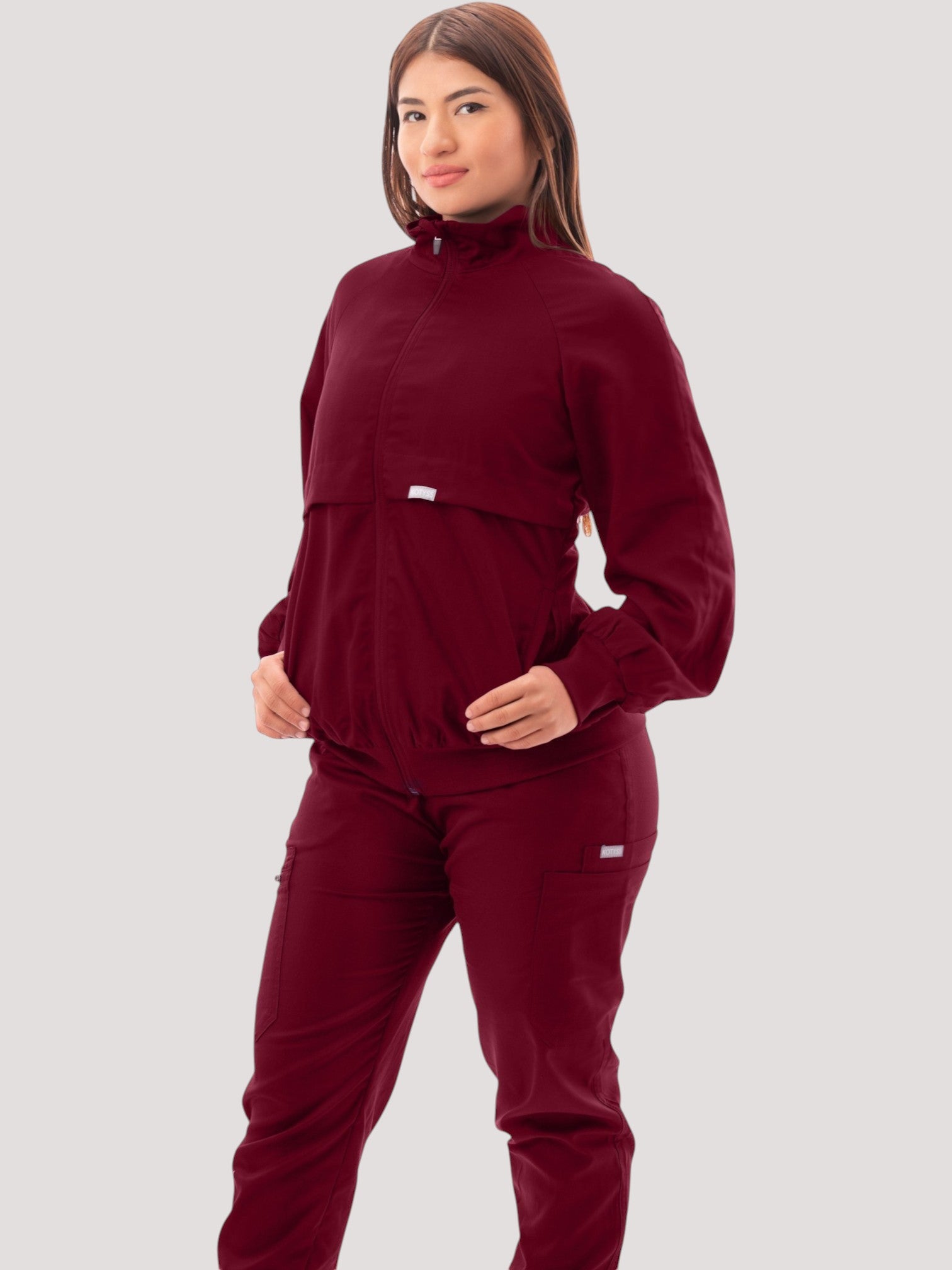 A model wearing a burgundy Kotyss women's scrub jacket with a high windbreaker neck and ribbed cuffs.