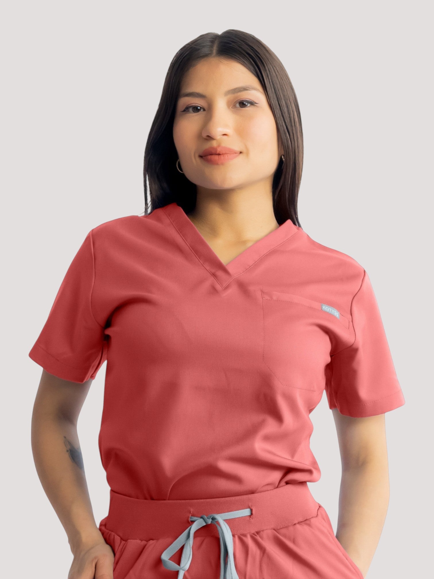 Woman wearing Kotyss coral pink V-neck scrub top with short sleeves and chest pocket, soft breathable fabric.