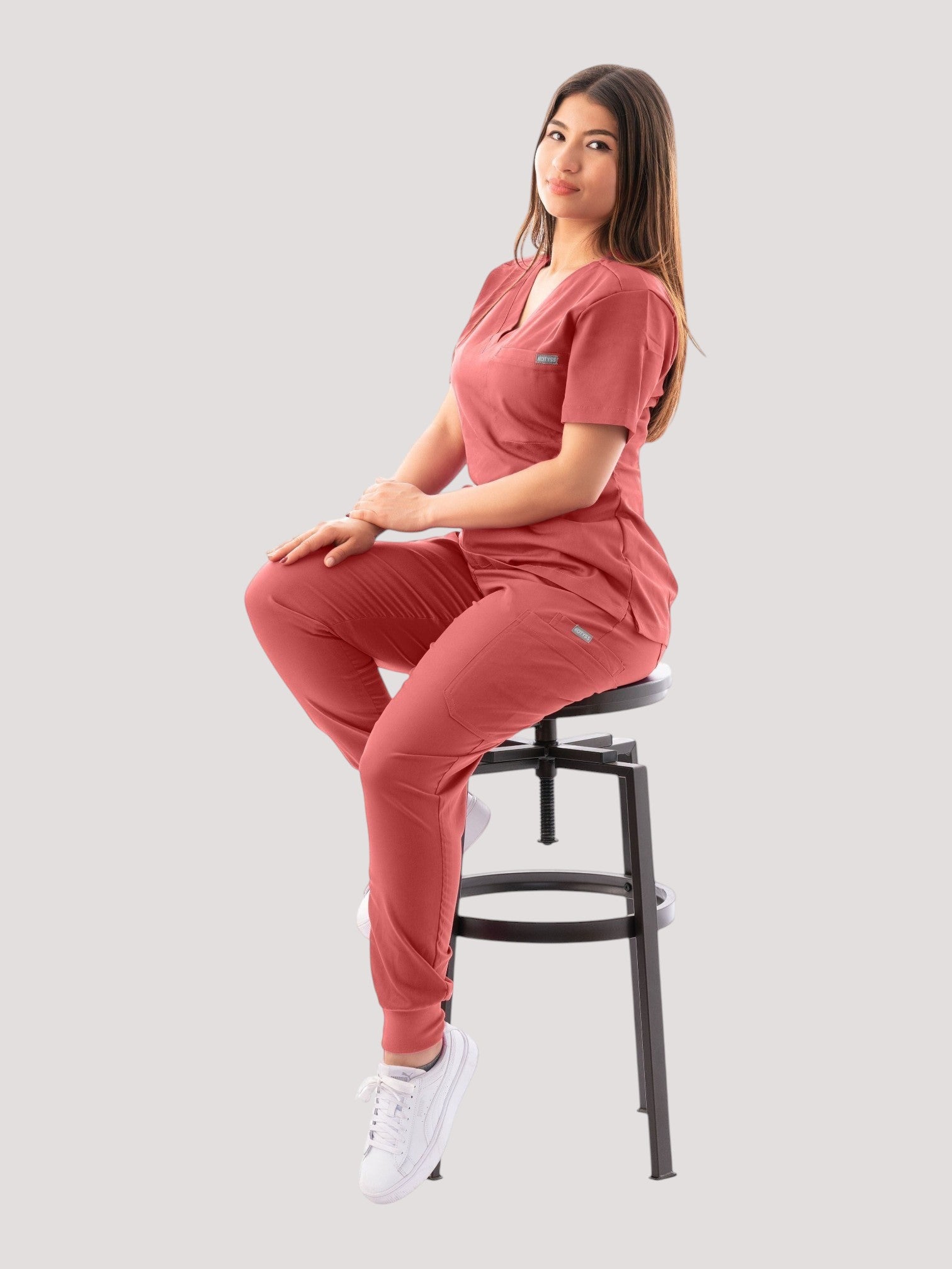 A woman wearing a Kotyss coral V-neck scrub top and jogger pants set, sitting on a stool against a white background.