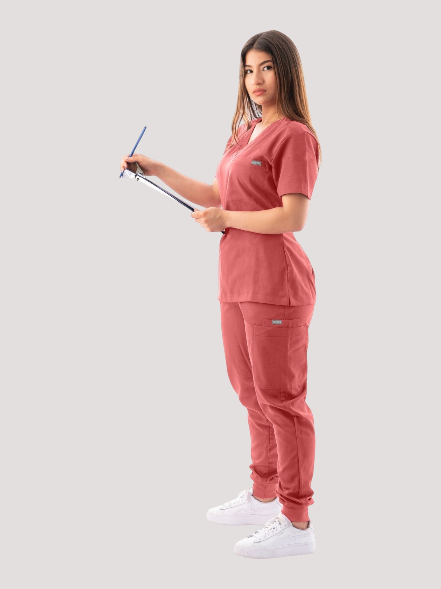 Kotyss women's coral V-neck scrub top and jogger pants set worn by a model holding a clipboard on a white background