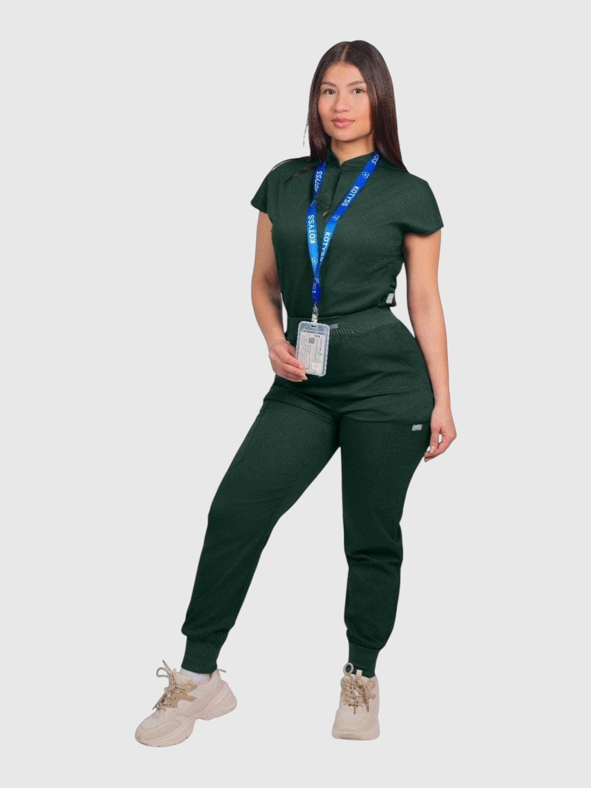 A woman wearing a dark green Kotyss collared scrub top and matching jogger pants with a branded lanyard.