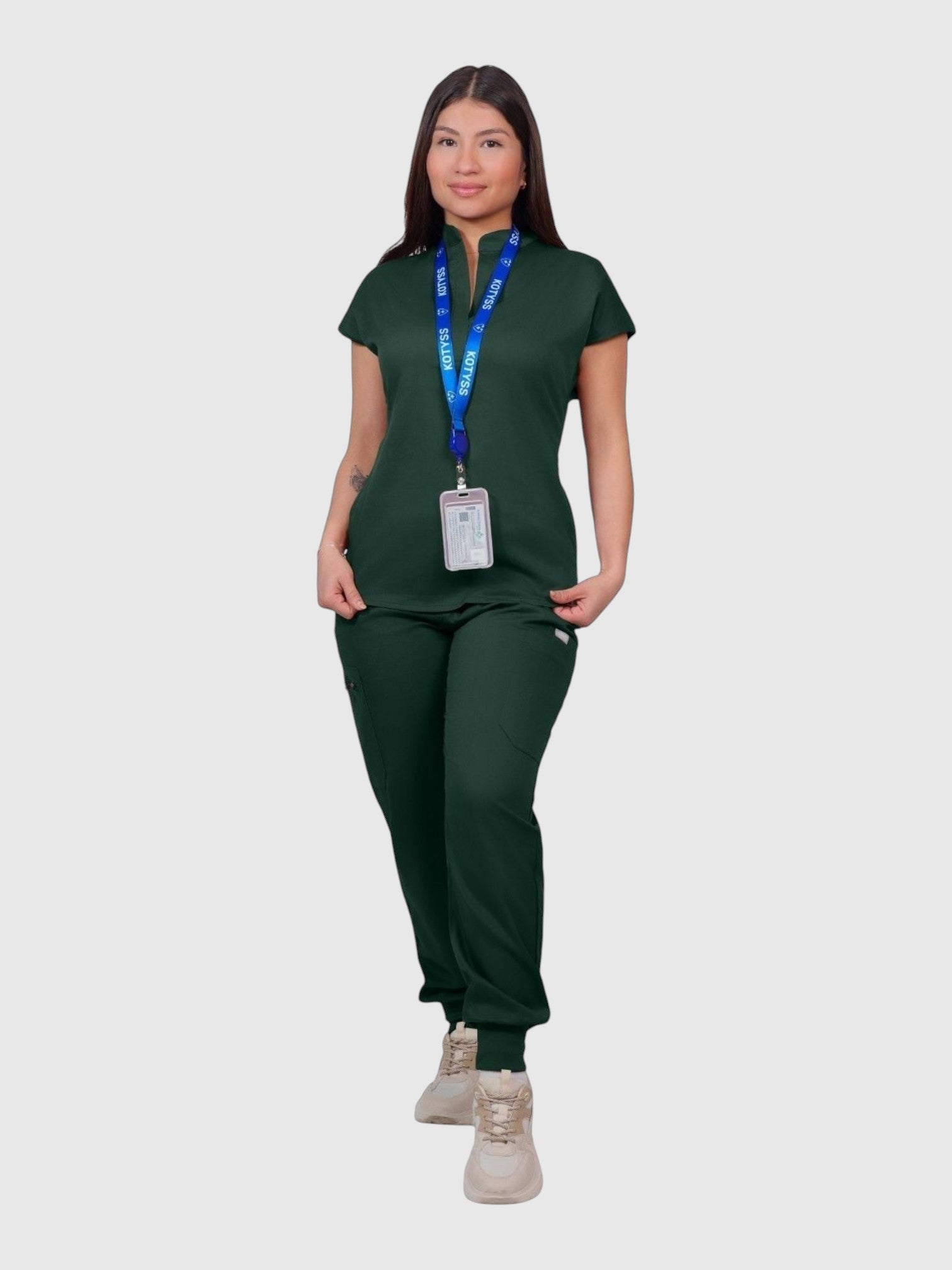 A woman wearing a dark green Kotyss collared scrub top and matching jogger pants with a branded lanyard.