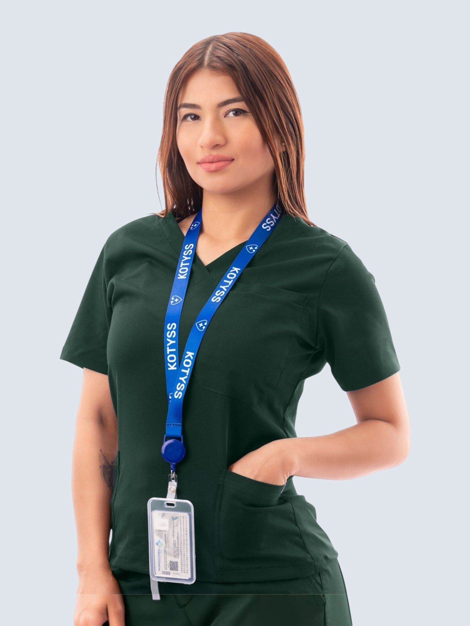 A woman wearing a dark green Kotyss V-neck medical scrub top with multiple pockets and a branded blue lanyard.