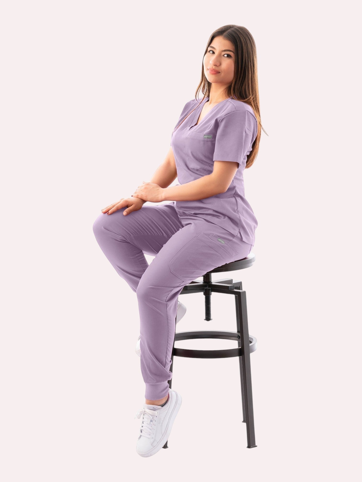 A woman wearing a lavender Kotyss V-neck scrub top and matching jogger pants, sitting on a stool.
