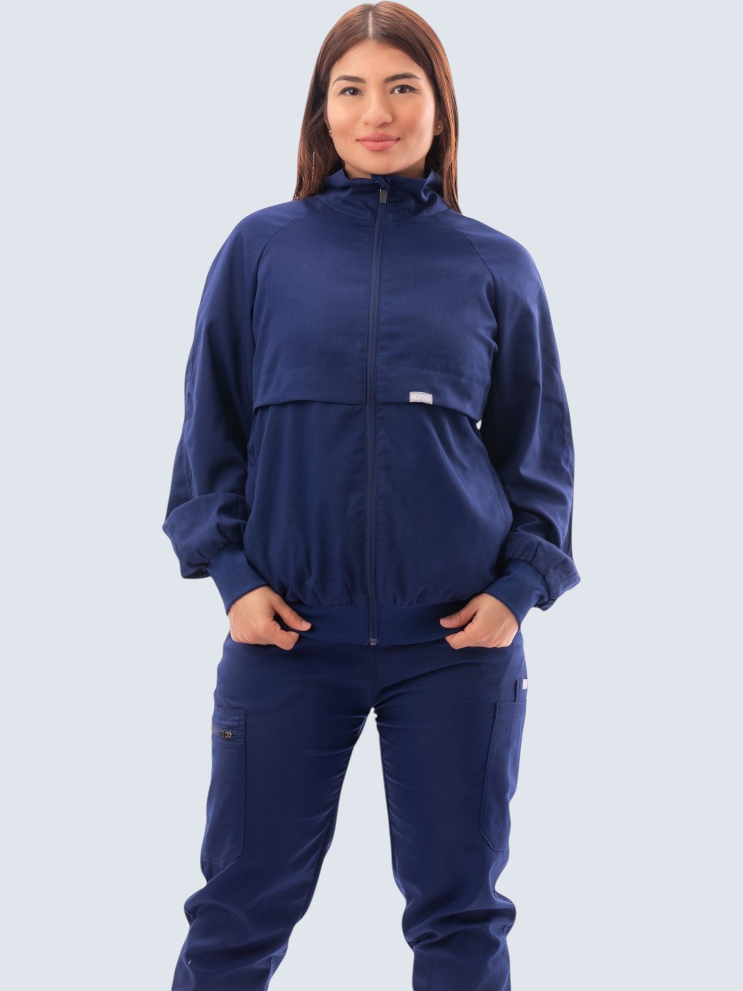 A woman wearing a navy blue Kotyss scrub jacket with a high windbreaker neck and ribbed cuffs on a white background.
