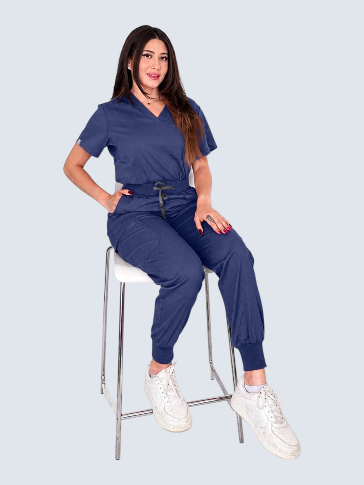 Kotyss navy blue V-neck scrub top and jogger pants set worn by a woman sitting on a white stool.