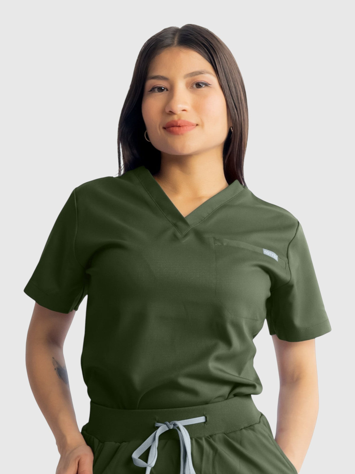 A woman wearing a dark olive green Kotyss V-neck scrub top with a chest pocket, featuring breathable stretch fabric.