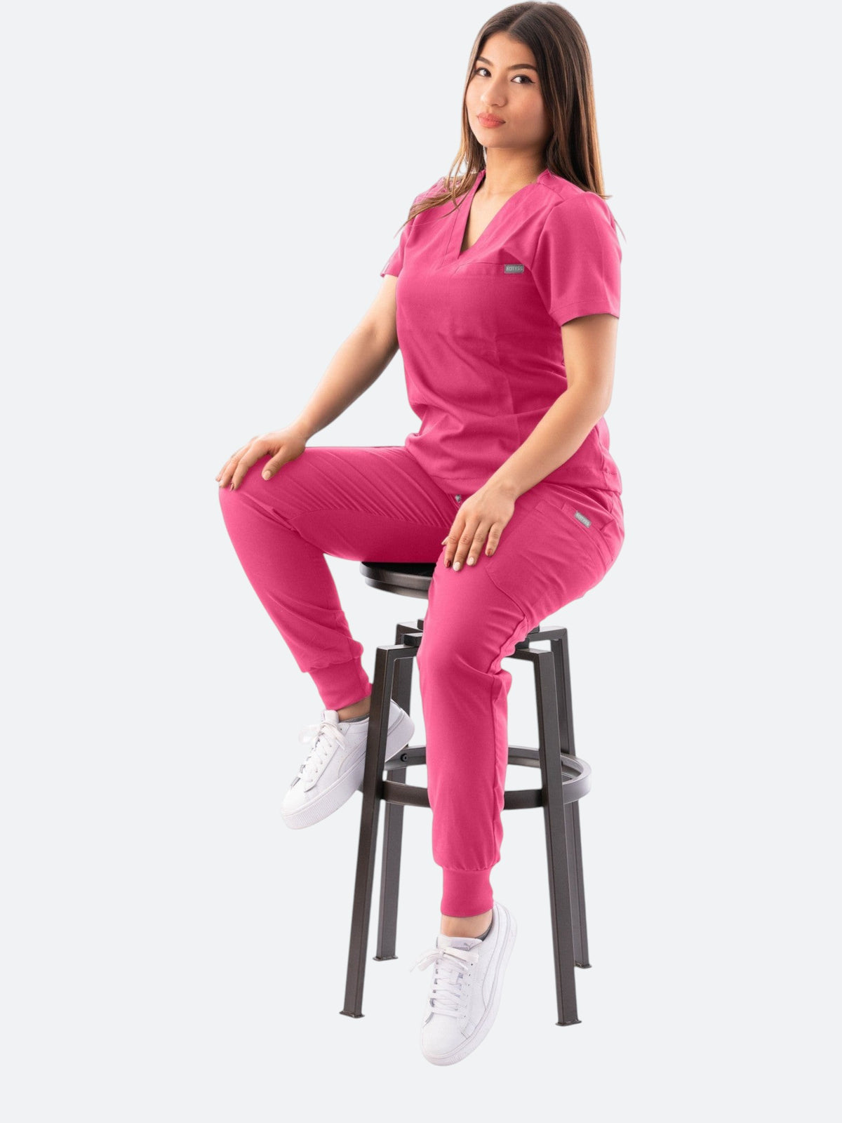 Model wearing a pink Kotyss V-neck scrub top and jogger pants set with multiple pockets, sitting on a stool.
