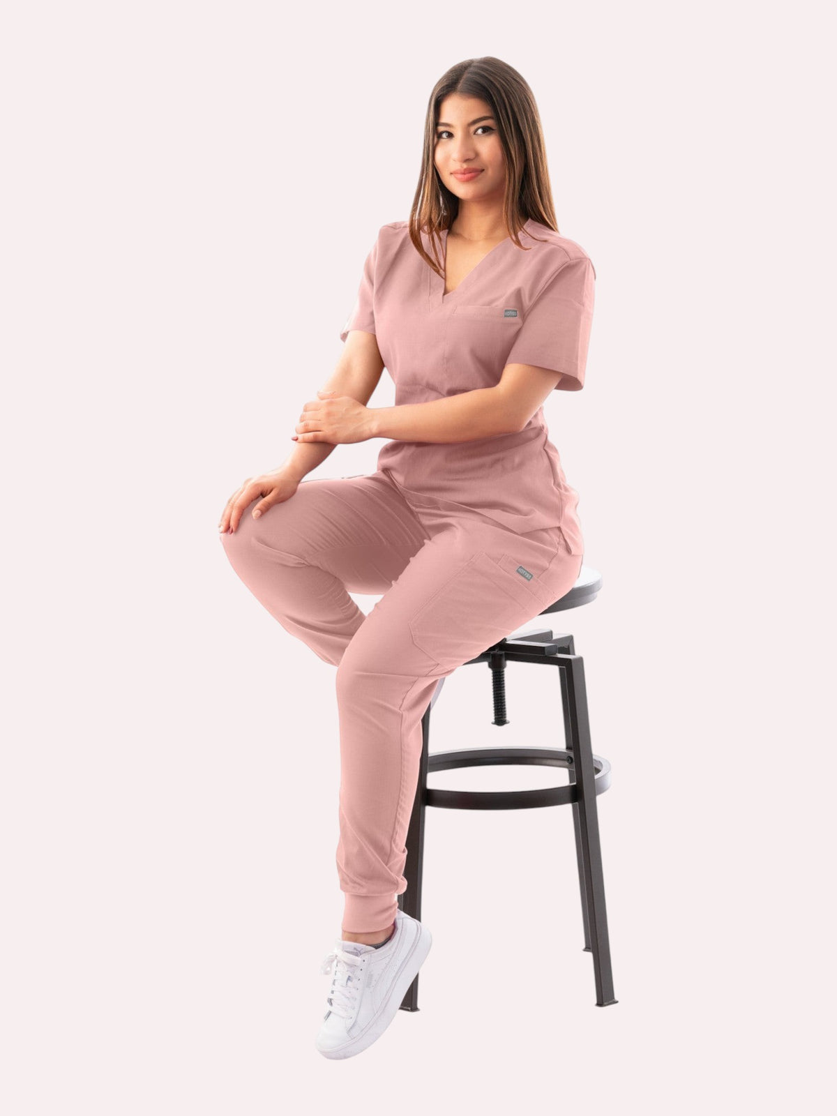 A woman wearing a dusty pink Kotyss V-neck scrub top and jogger pants set while sitting on a stool.