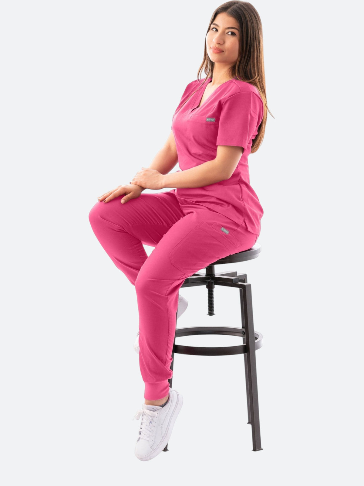 A woman wearing a pink Kotyss V-neck scrub top and jogger pants set, sitting on a stool against a white background.