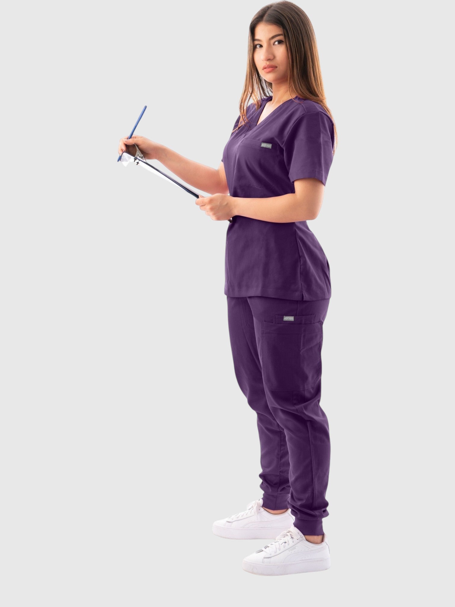 Kotyss purple V-neck scrub top and jogger pants set worn by a woman holding a clipboard