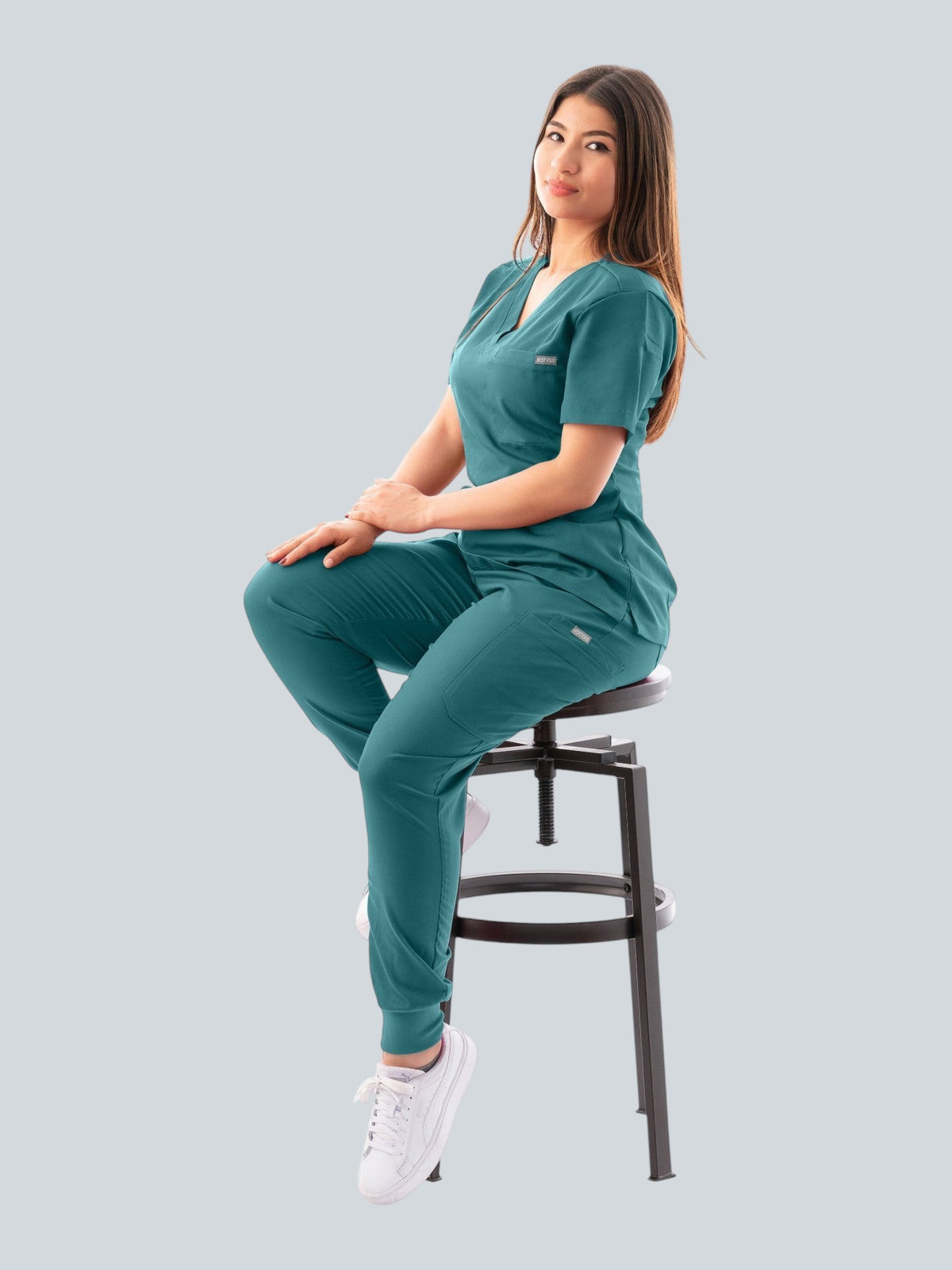 Woman wearing a teal Kotyss V-neck scrub top and jogger pants set while sitting on a stool