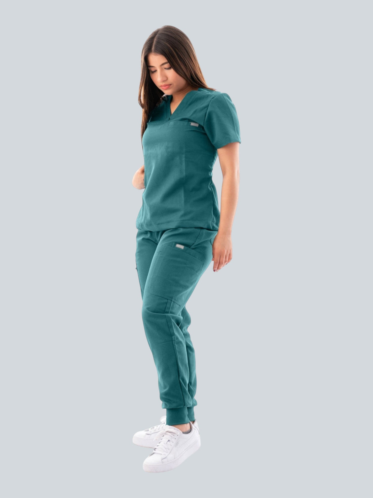 A woman wearing a teal Kotyss V-neck scrub top and matching jogger pants with multiple pockets and white sneakers.