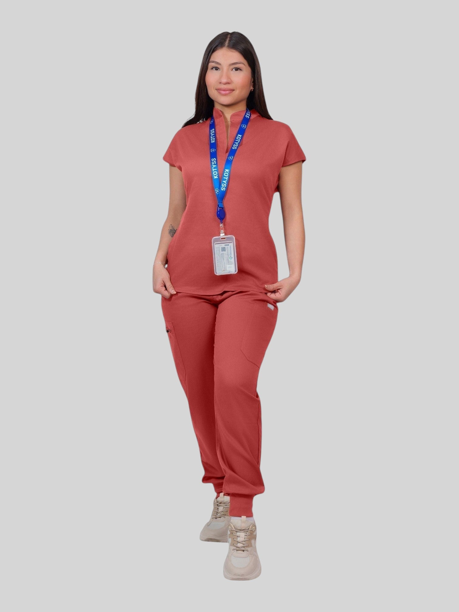 A woman wearing a Kotyss terracotta collared scrub top and matching jogger pants with a blue branded lanyard.