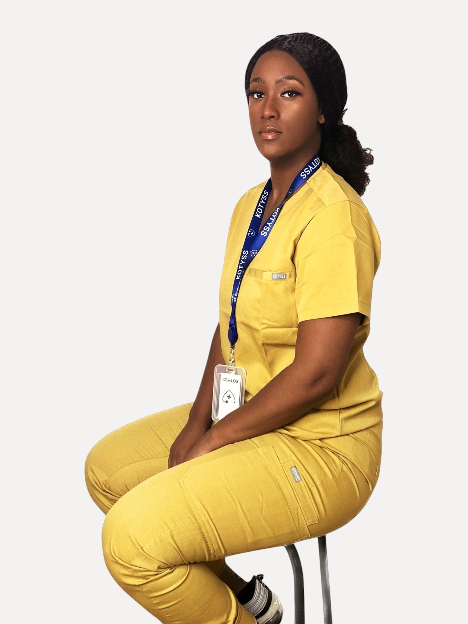 A woman wearing a yellow Kotyss V-neck scrub top and jogger pants set with multiple pockets, sitting on a stool.