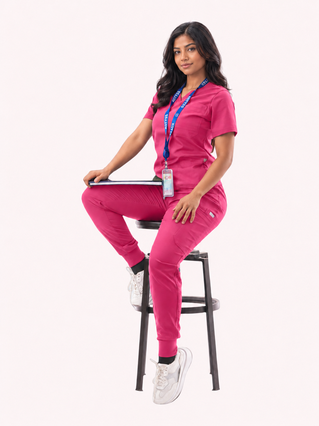 A woman wearing a matching bright pink medical scrub set sits on a black stool against a light pink background. She is holding a laptop and wearing a blue lanyard with an ID badge.