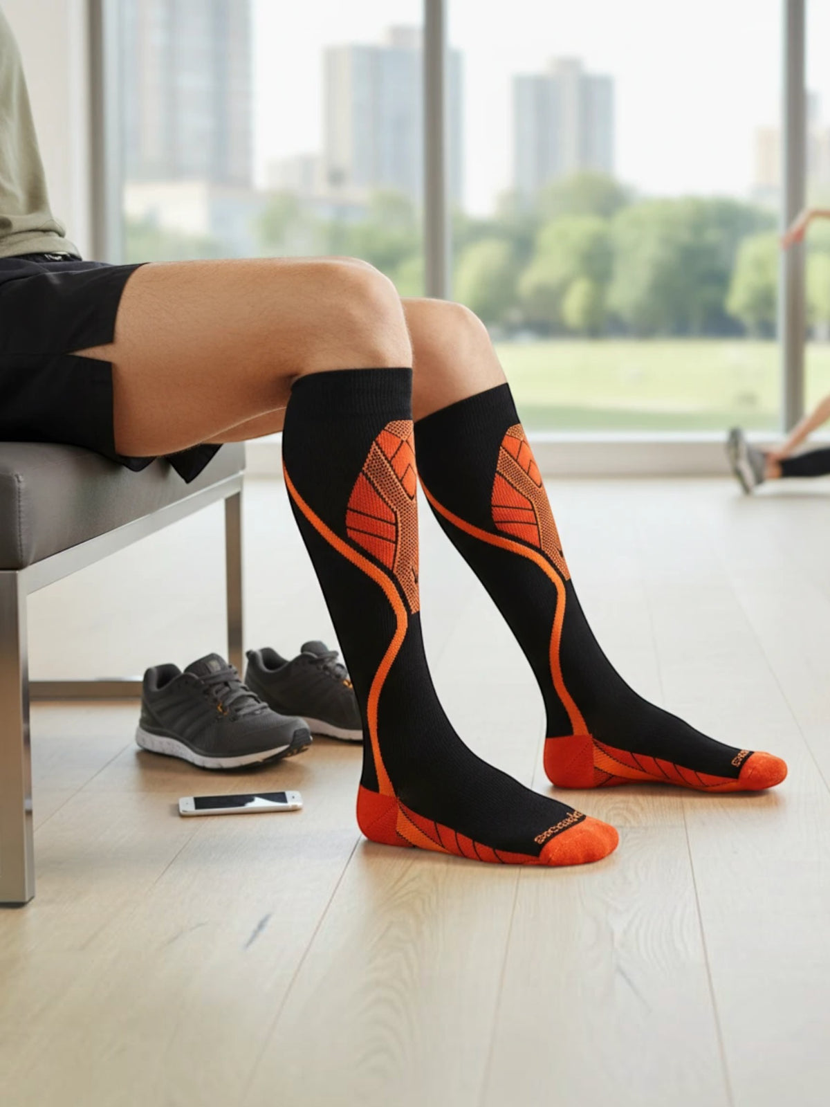 Kotyss knee-high compression socks in black and orange with athletic pattern, worn by a person sitting on a bench.