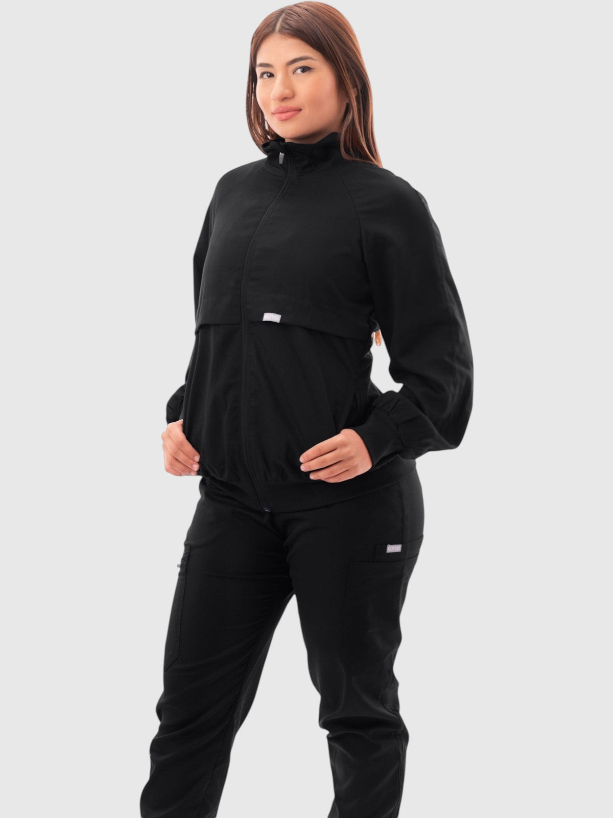 A woman wearing a black Kotyss scrub jacket with a high windbreaker neck, ribbed cuffs, and functional pockets.