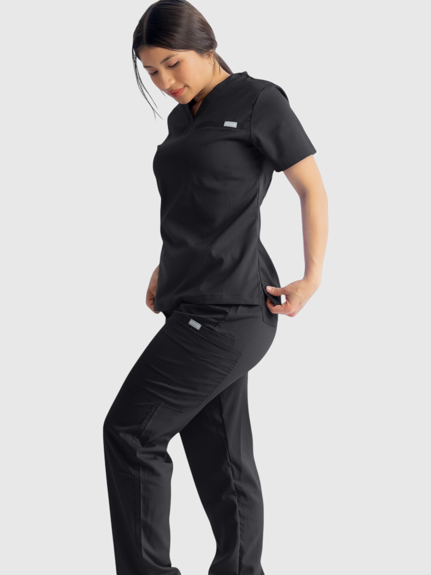 Kotyss black straight cut scrub pants and matching top worn by a woman, featuring 8 pockets and 4-way stretch fabric.