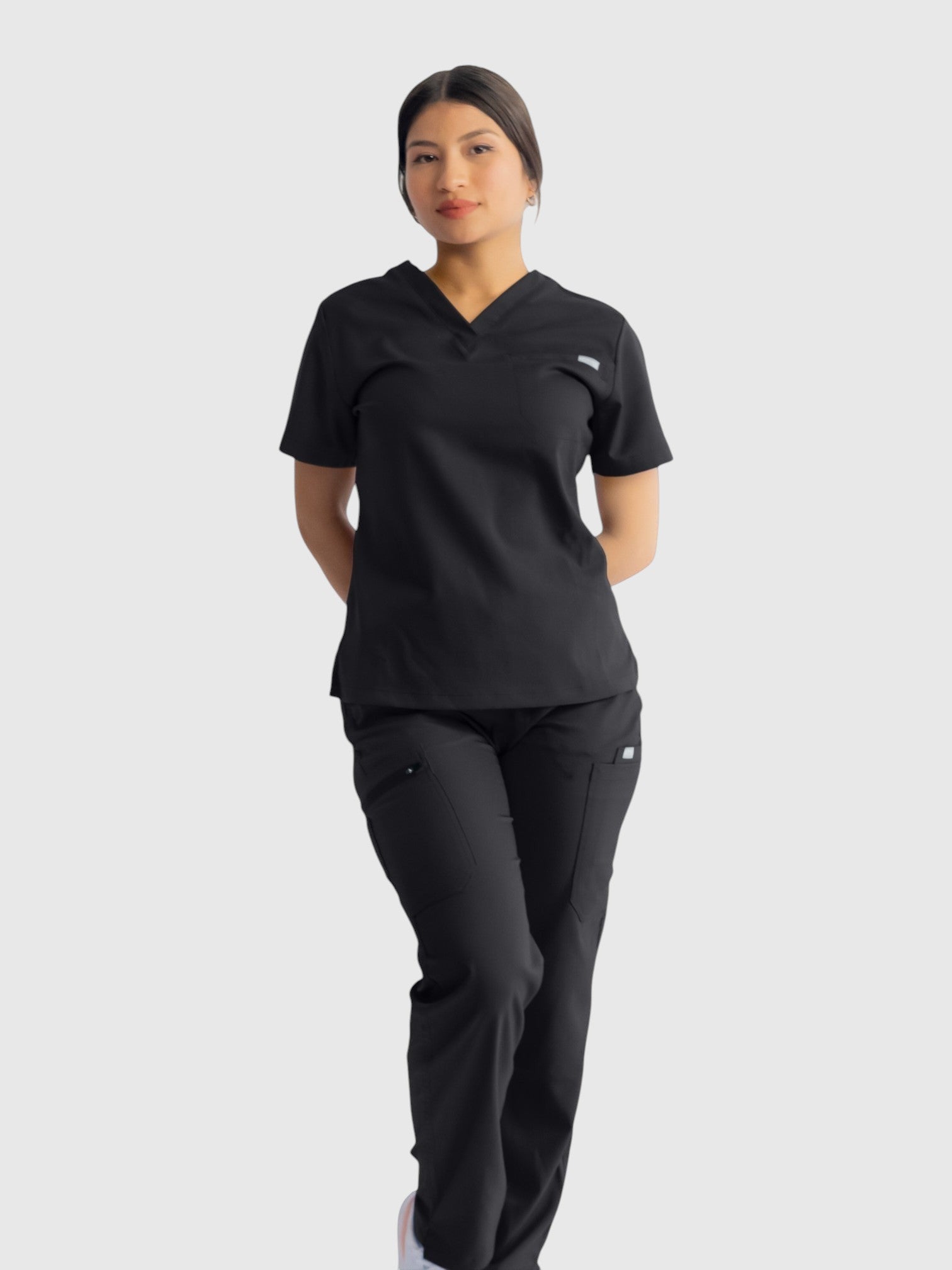 A woman wearing Kotyss black straight cut scrub pants and a matching V-neck scrub top, featuring 8 functional pockets.