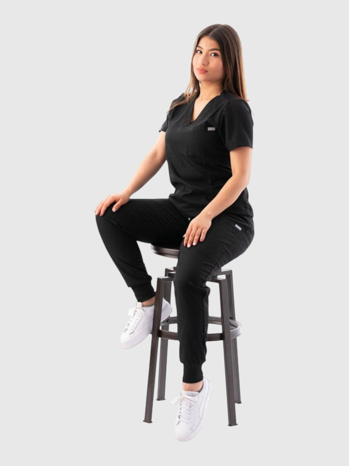 A woman wearing a black Kotyss V-neck scrub top and jogger pants, sitting on a stool, showing a professional fit.