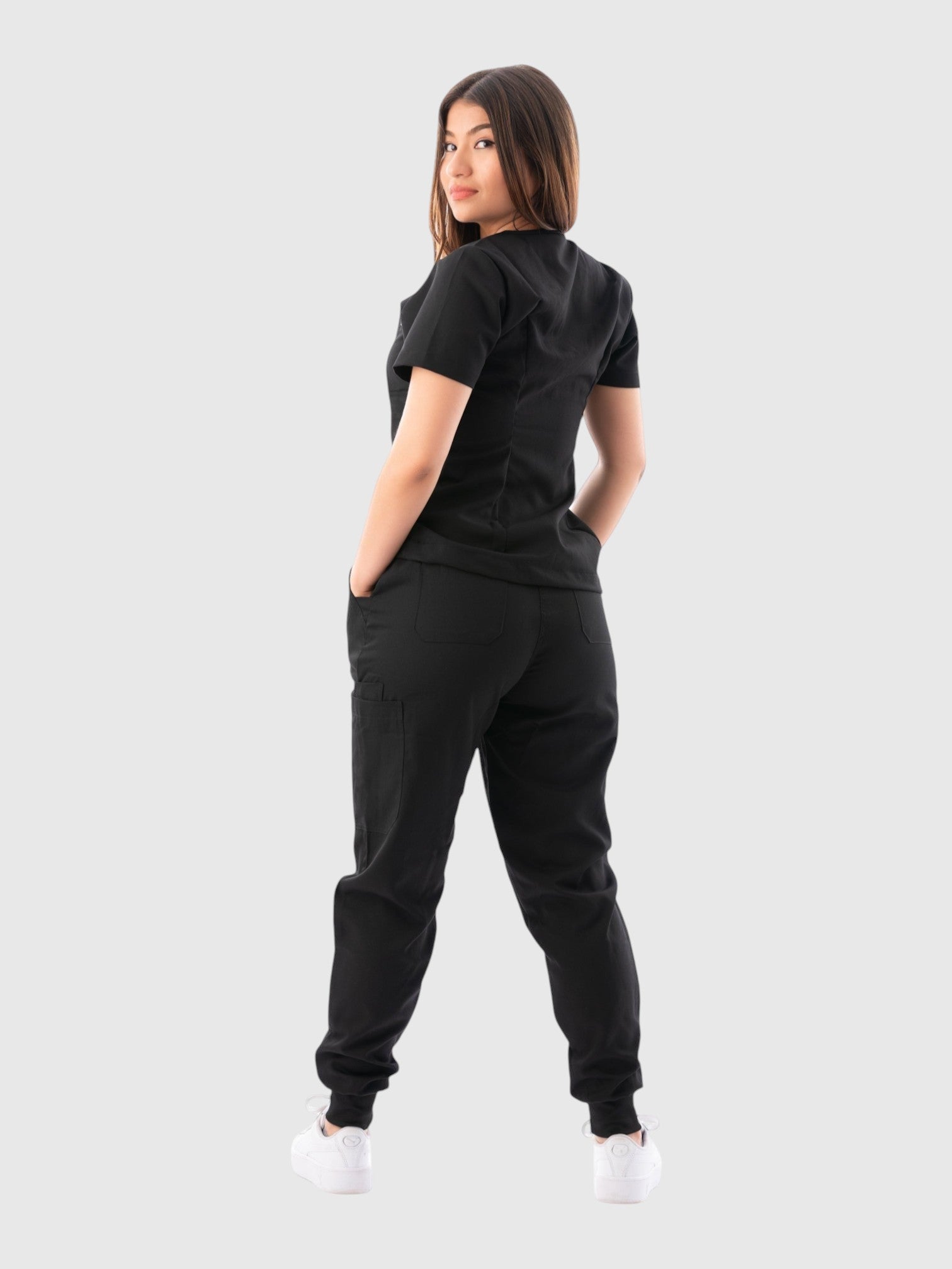 A woman wearing a black Kotyss V-neck scrub top and jogger pants set, viewed from the back against a grey background.