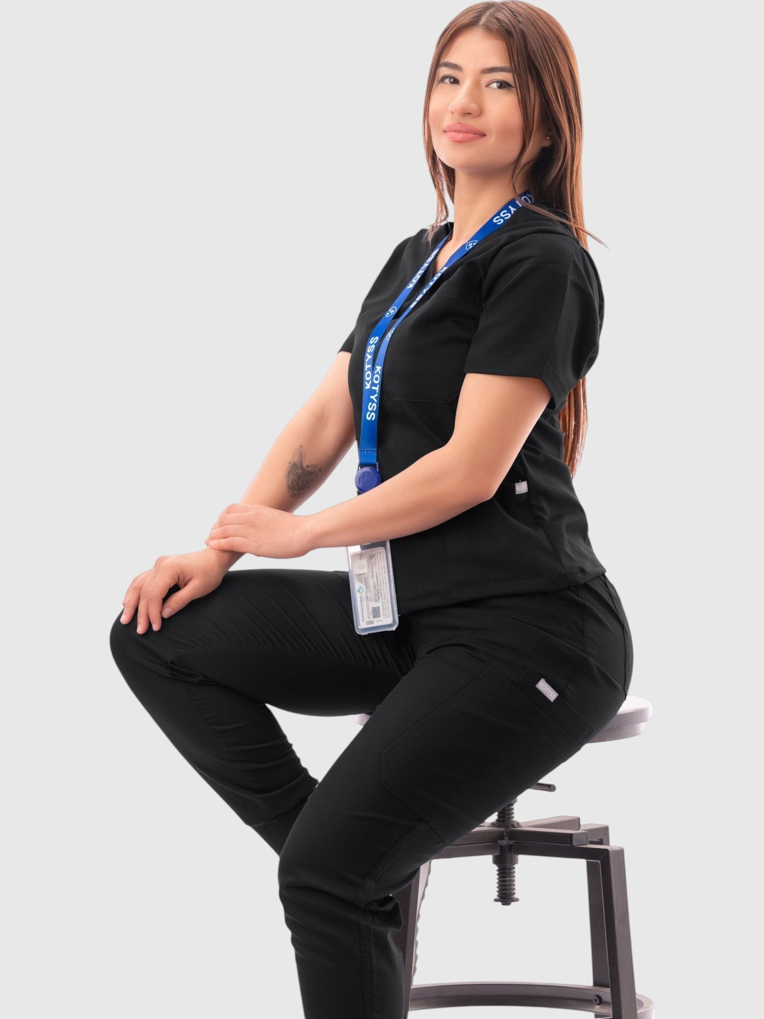 A woman wearing a black Kotyss V-neck scrub top and jogger pants set, sitting on a stool against a white background.