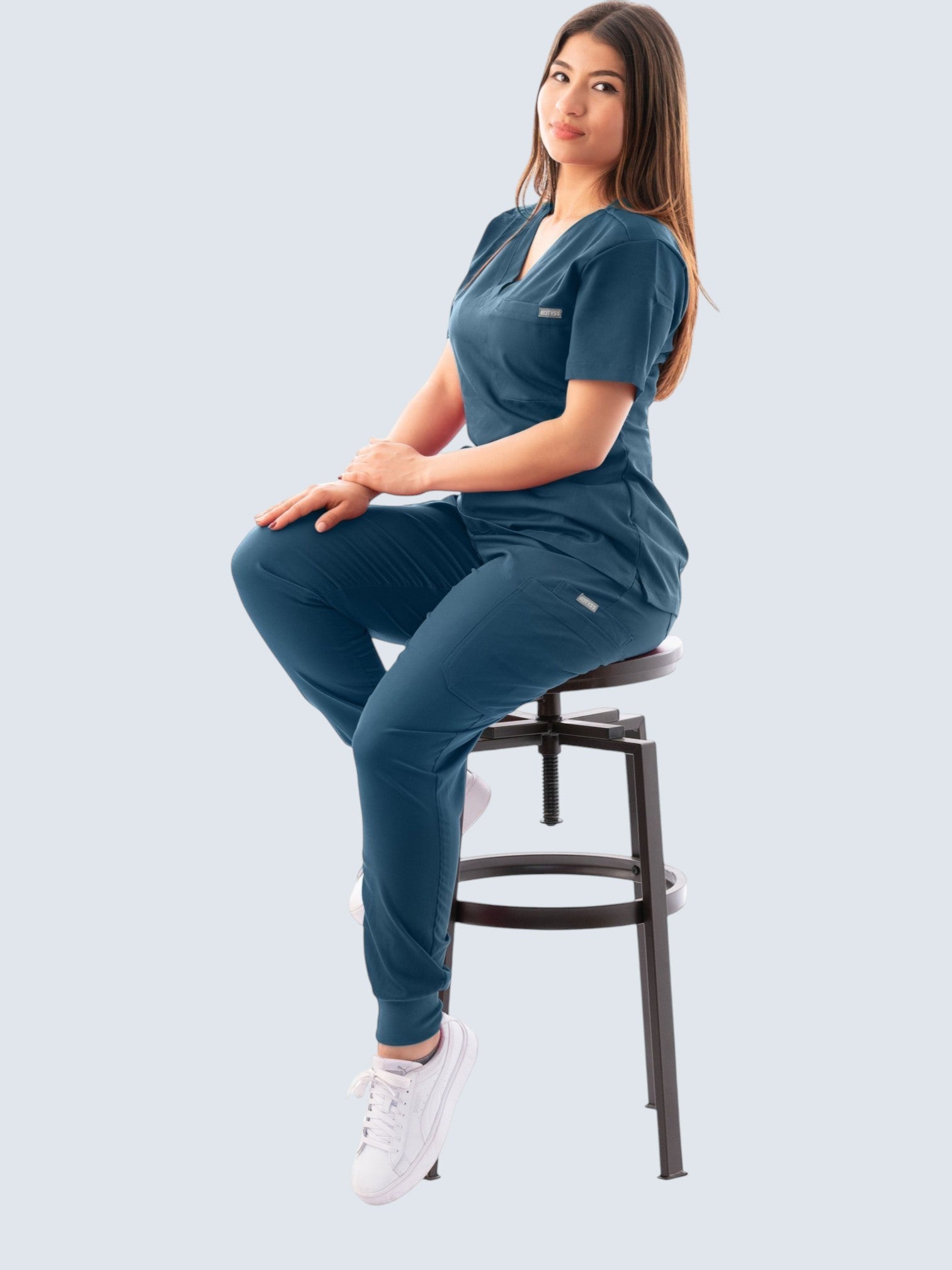 Woman wearing a dark blue Kotyss V-neck scrub top and jogger pants set while sitting on a stool.