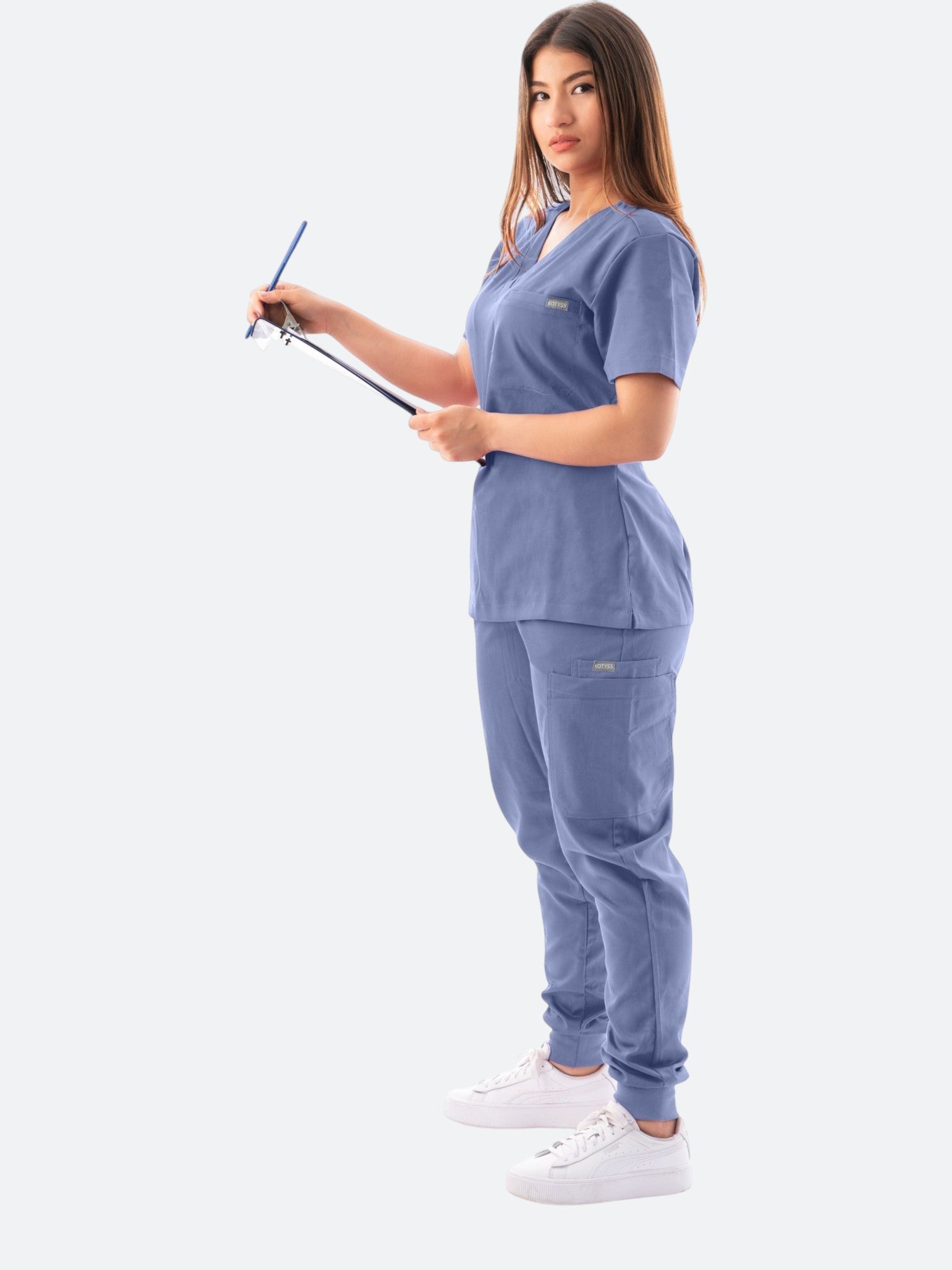 A woman wearing a blue Kotyss V-neck scrub top and jogger pants, holding a clipboard, standing against a white background.