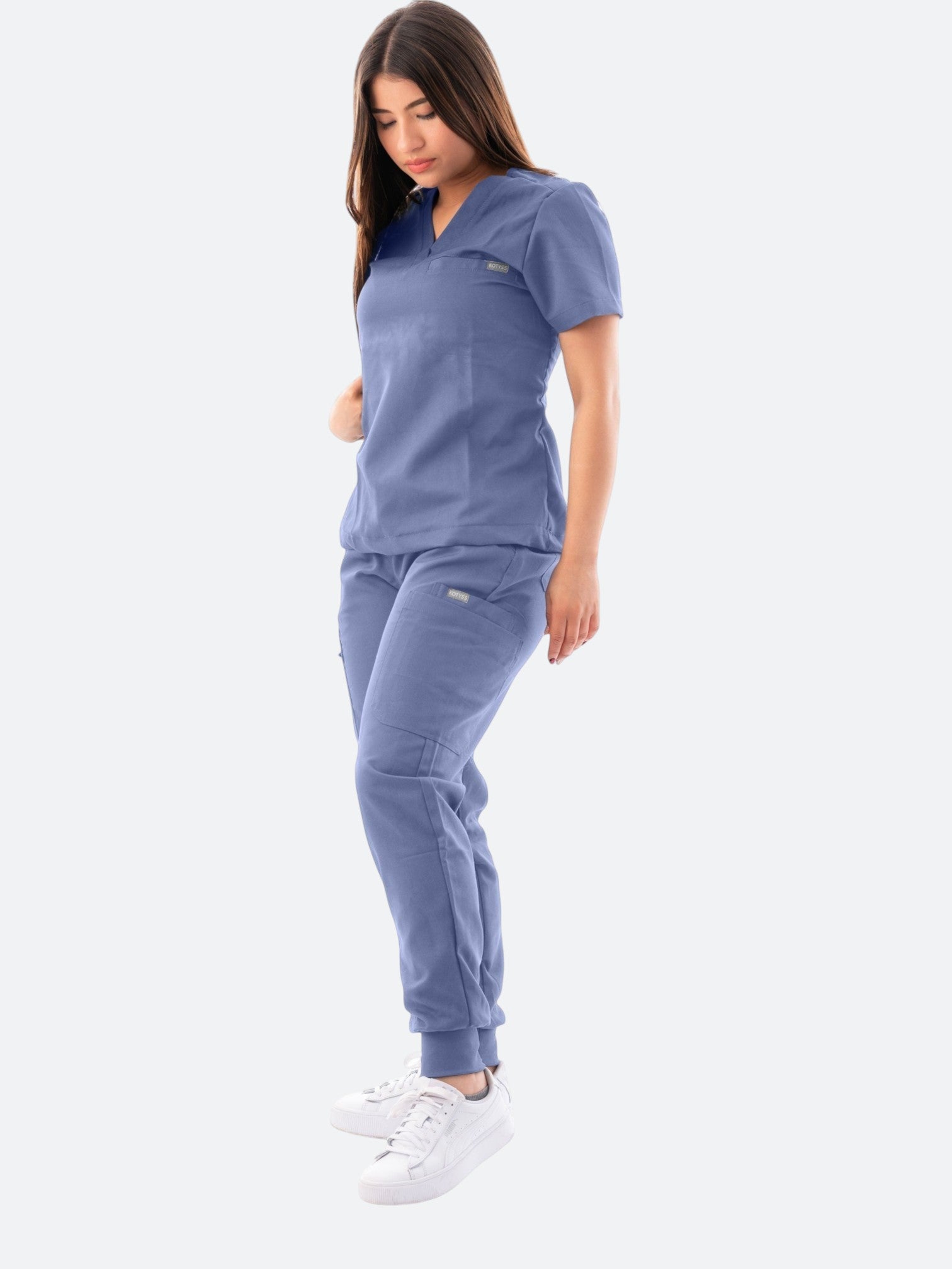 A woman wearing a blue Kotyss V-neck scrub top and matching jogger pants with multiple pockets.