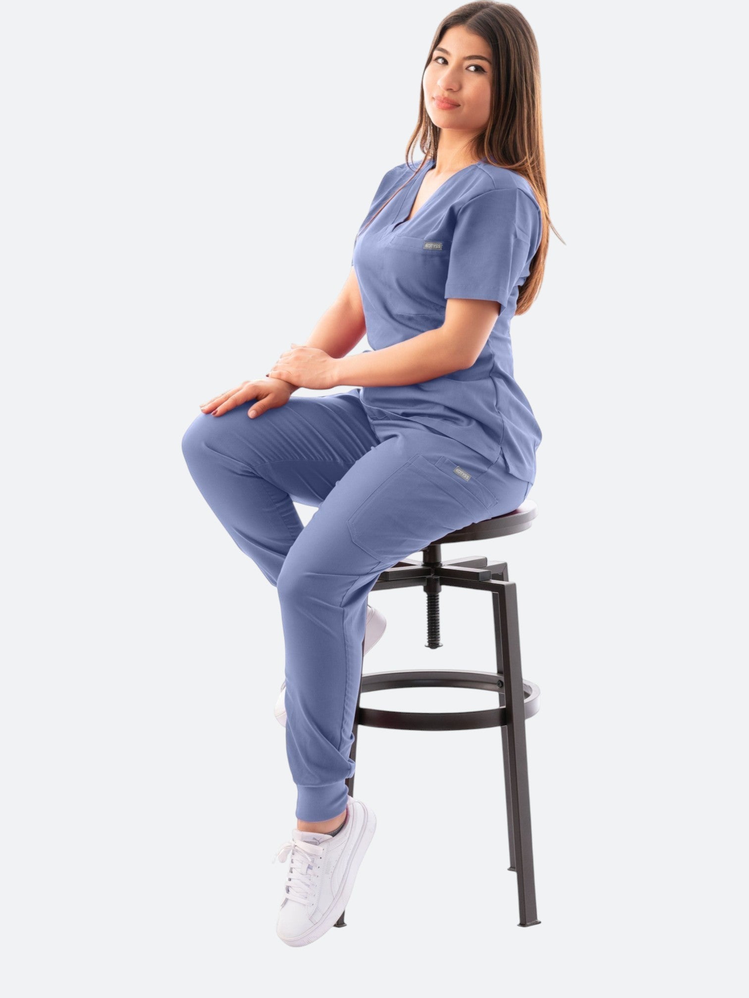 A woman wearing a blue Kotyss V-neck scrub top and matching jogger pants while sitting on a stool.
