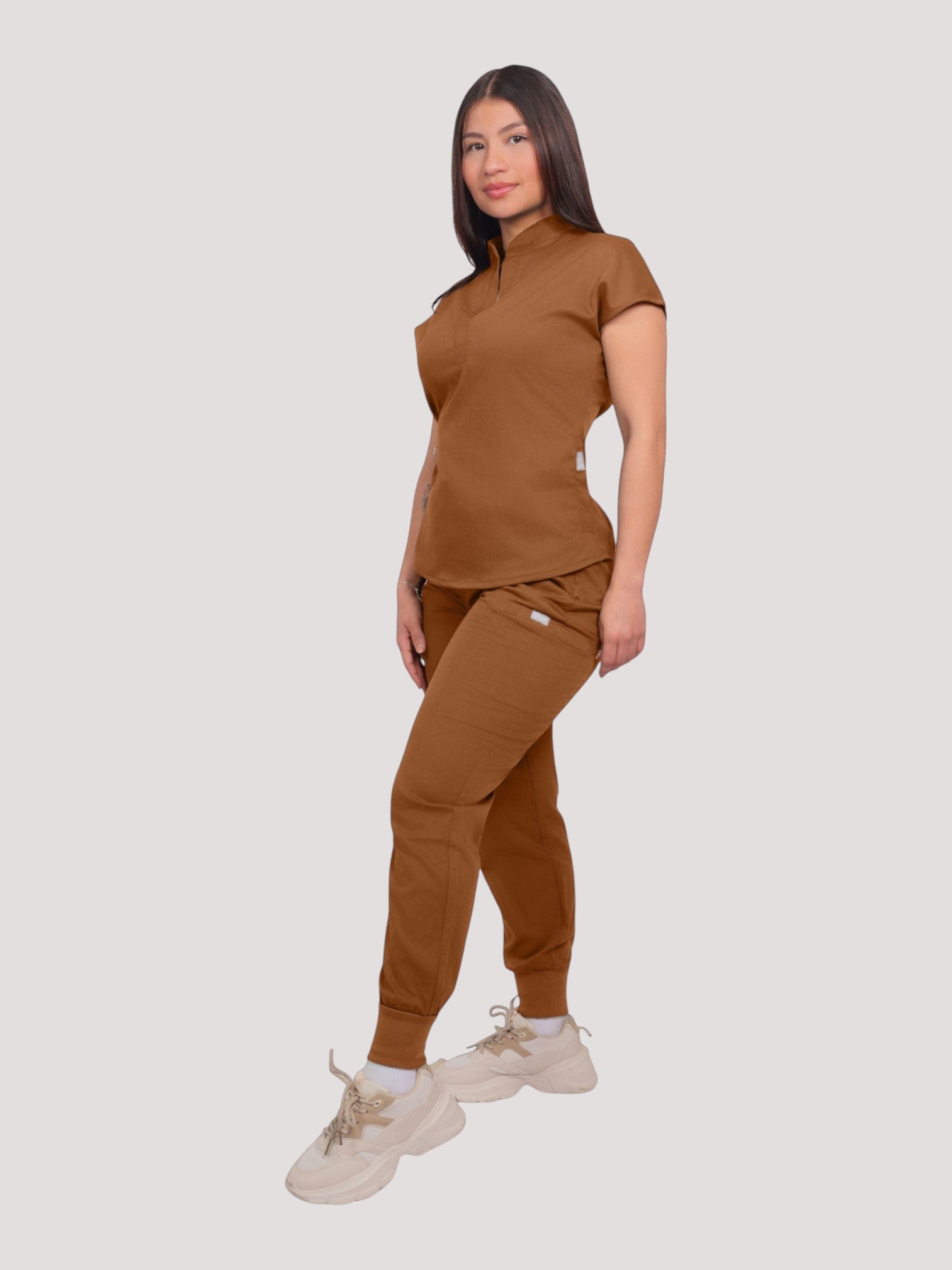A woman wearing a Kotyss brown collared scrub top and matching jogger pants with multiple pockets and 4-way stretch.