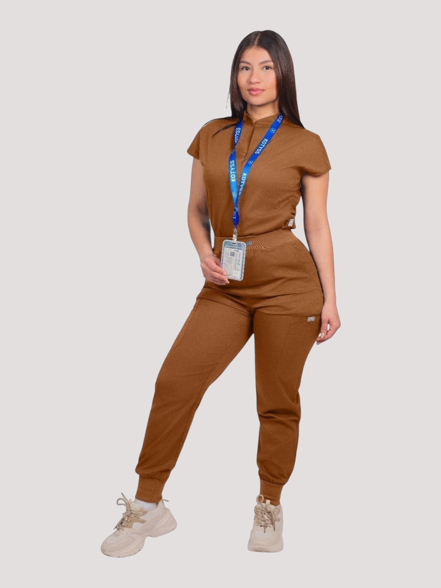 A woman wearing a brown Kotyss collared scrub top and matching jogger pants with a blue branded lanyard.