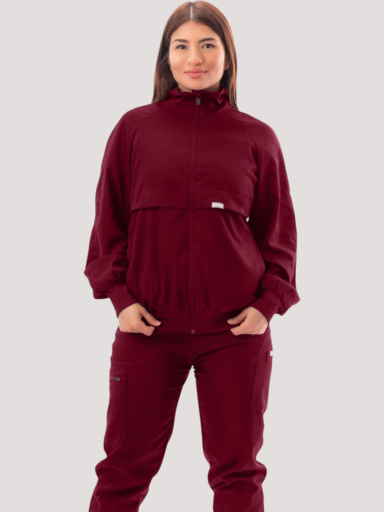 A woman wearing a Kotyss burgundy scrub jacket with a high windbreaker neck, ribbed cuffs, and a tailored silhouette.