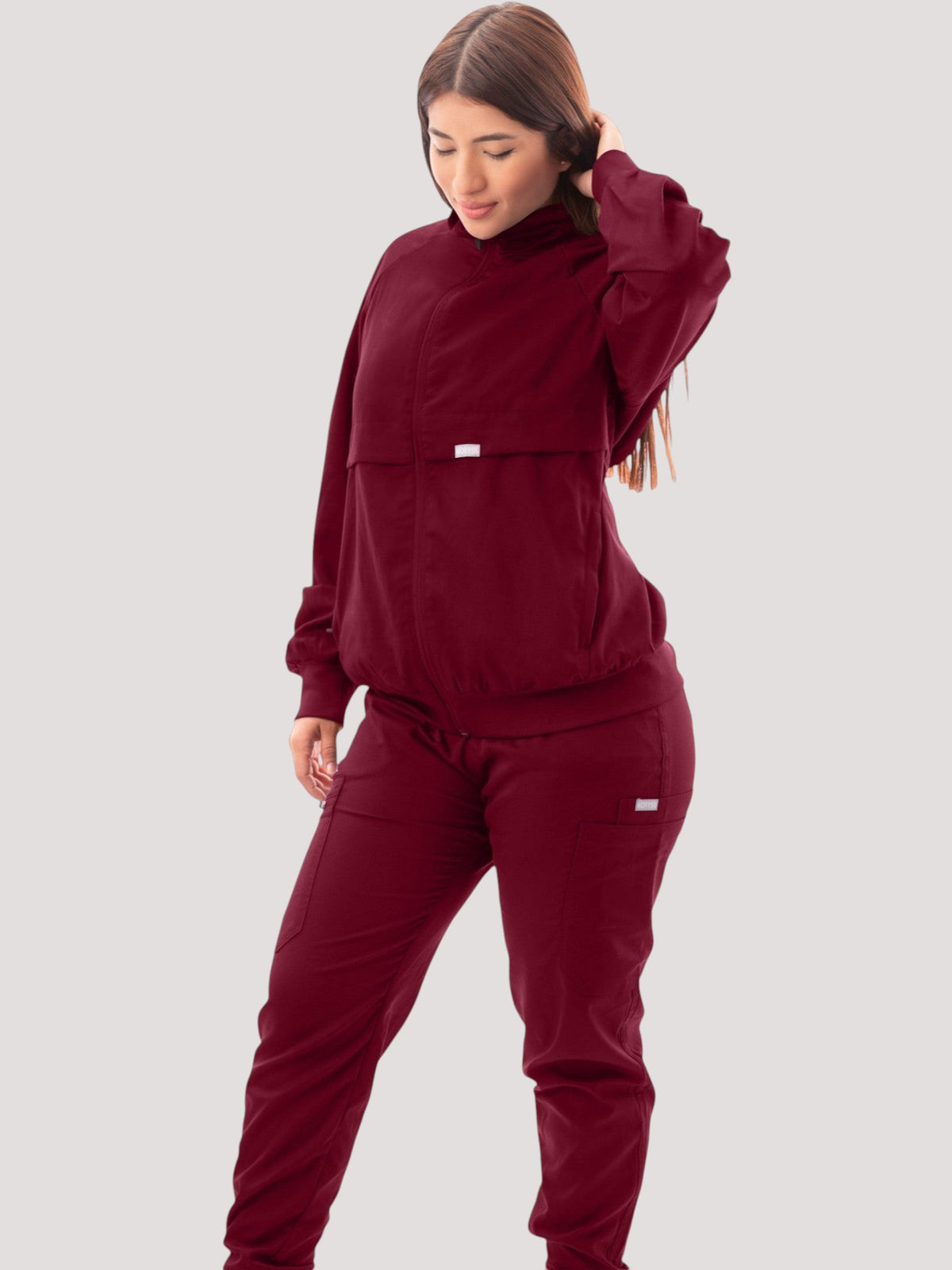 A woman wearing a Kotyss burgundy scrub jacket with a high neck, ribbed cuffs, and matching scrub pants.