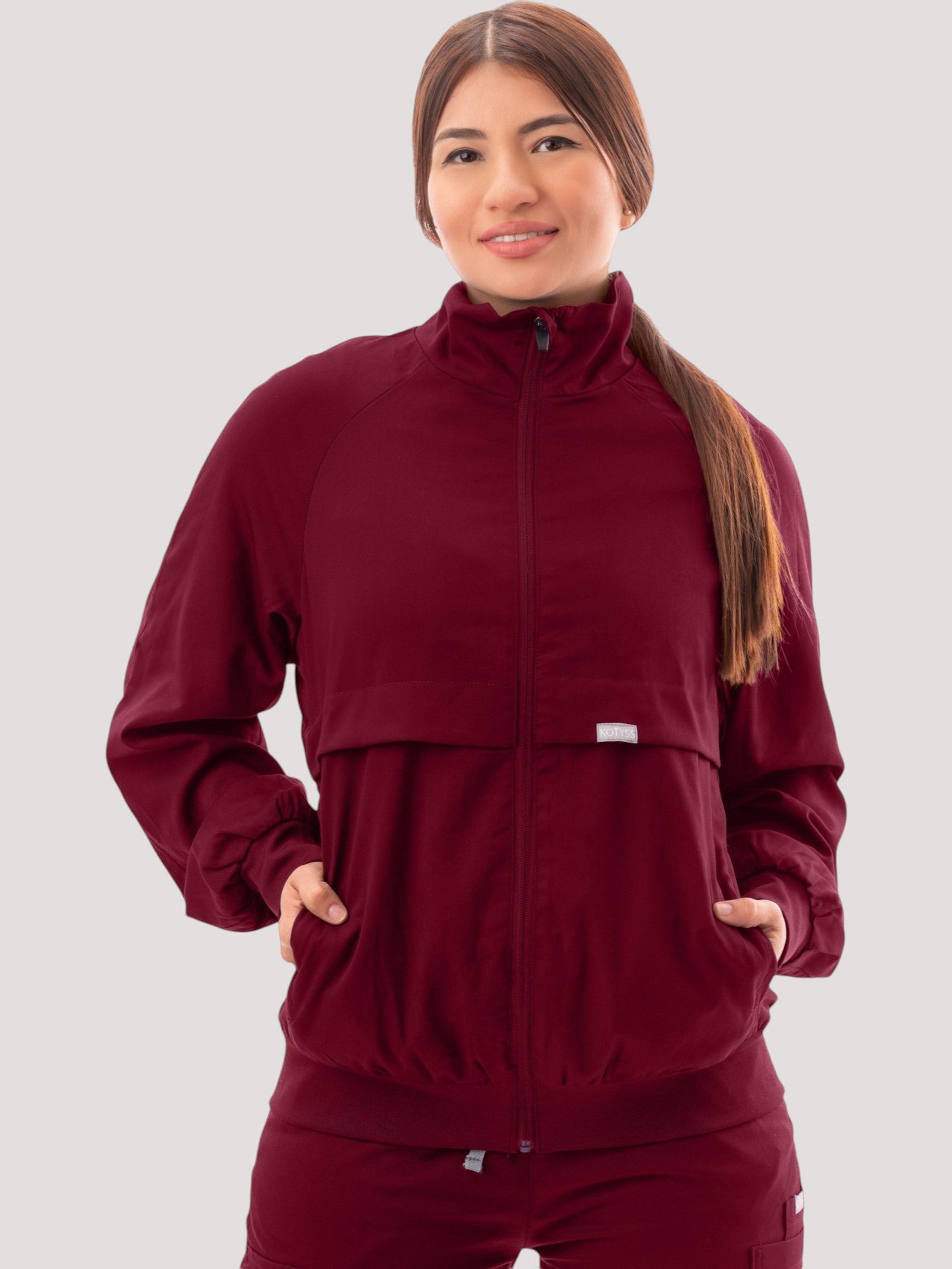 A woman wearing a burgundy Kotyss scrub jacket with a high windbreaker neck, ribbed cuffs, and functional pockets.
