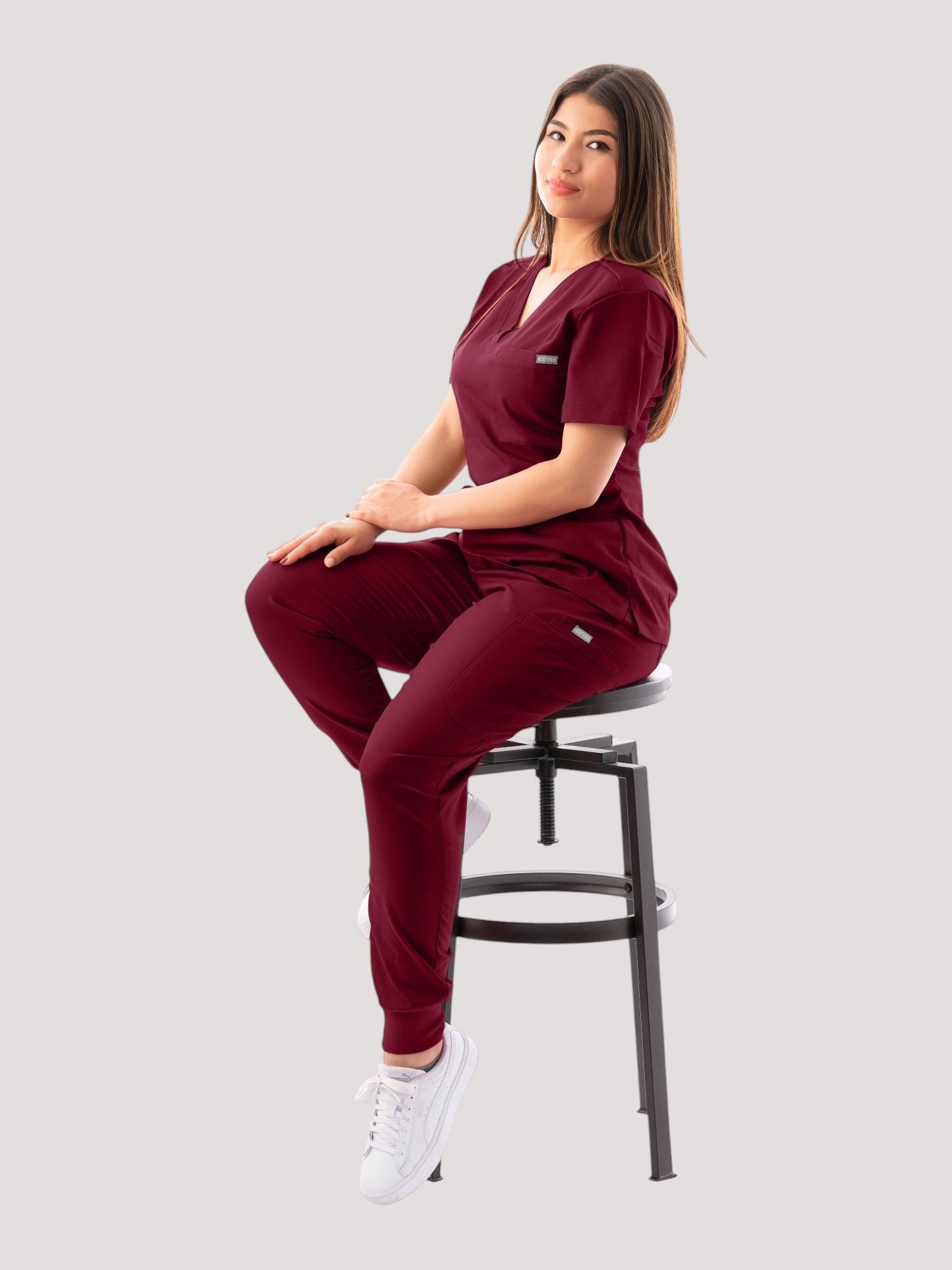 Woman wearing a Kotyss burgundy V-neck scrub top and jogger pants set while sitting on a stool