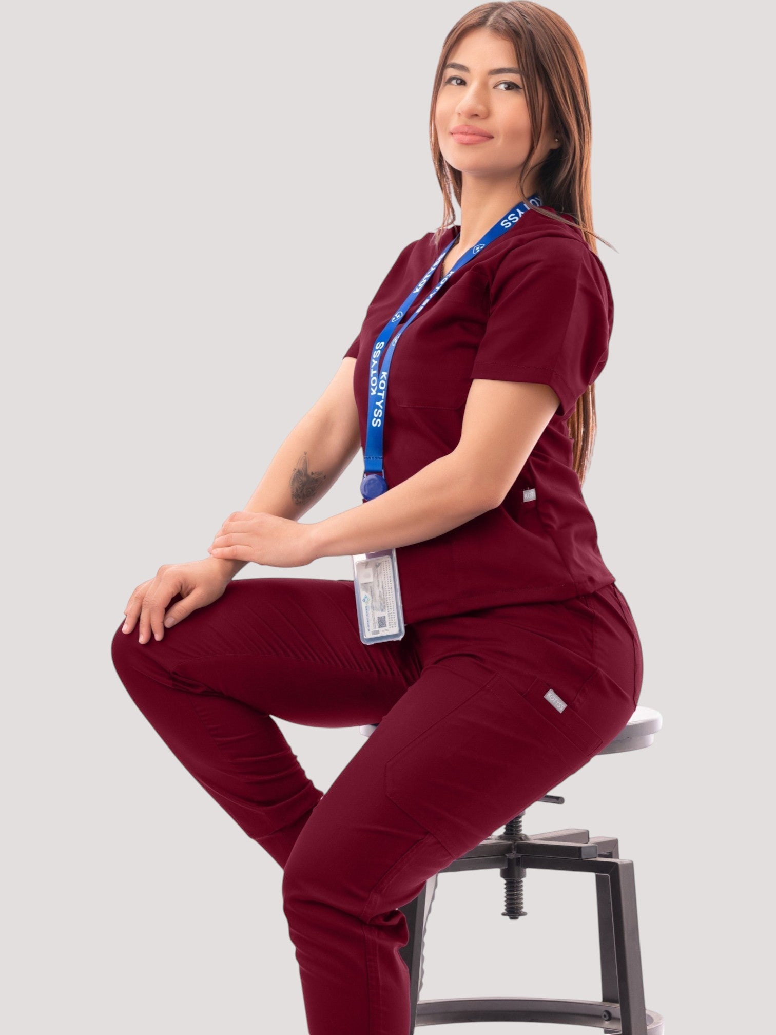 Kotyss burgundy V-neck scrub top and jogger pants set worn by a woman sitting on a stool