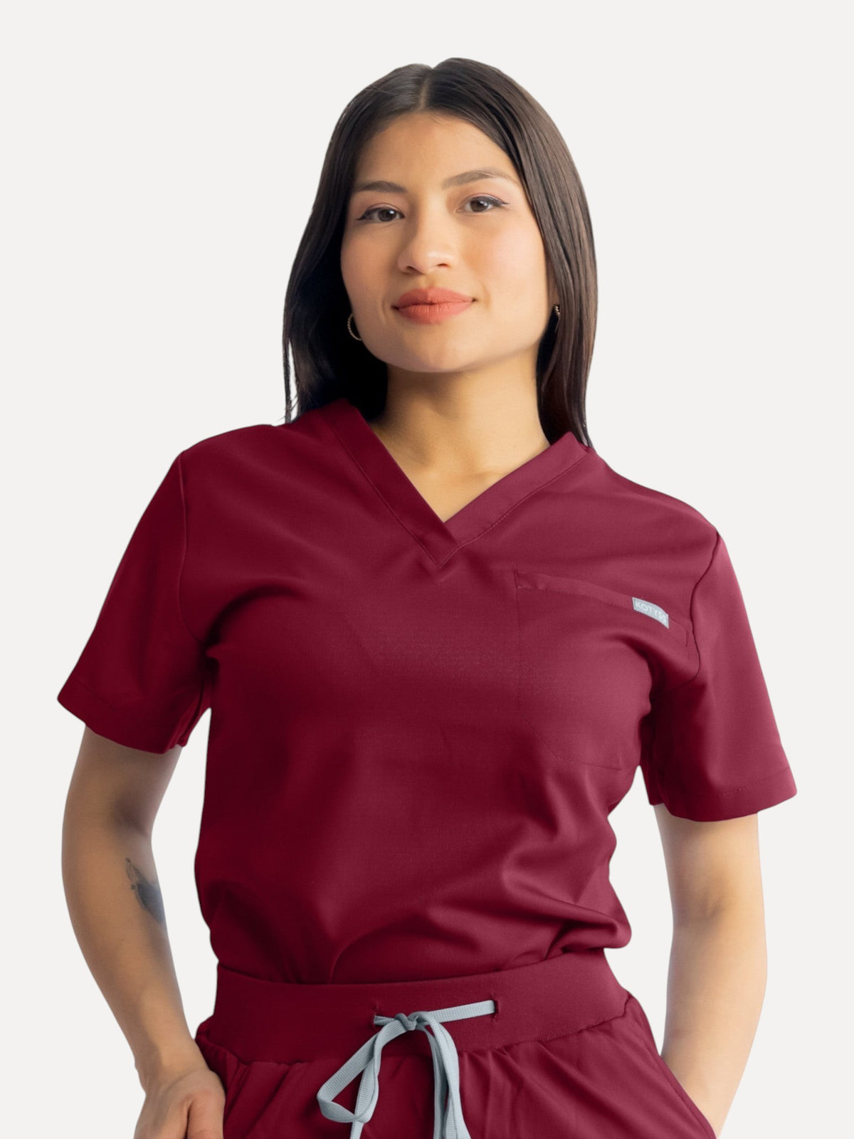 A woman wearing a burgundy Kotyss V-neck scrub top with a chest pocket, featuring a soft, breathable fabric.