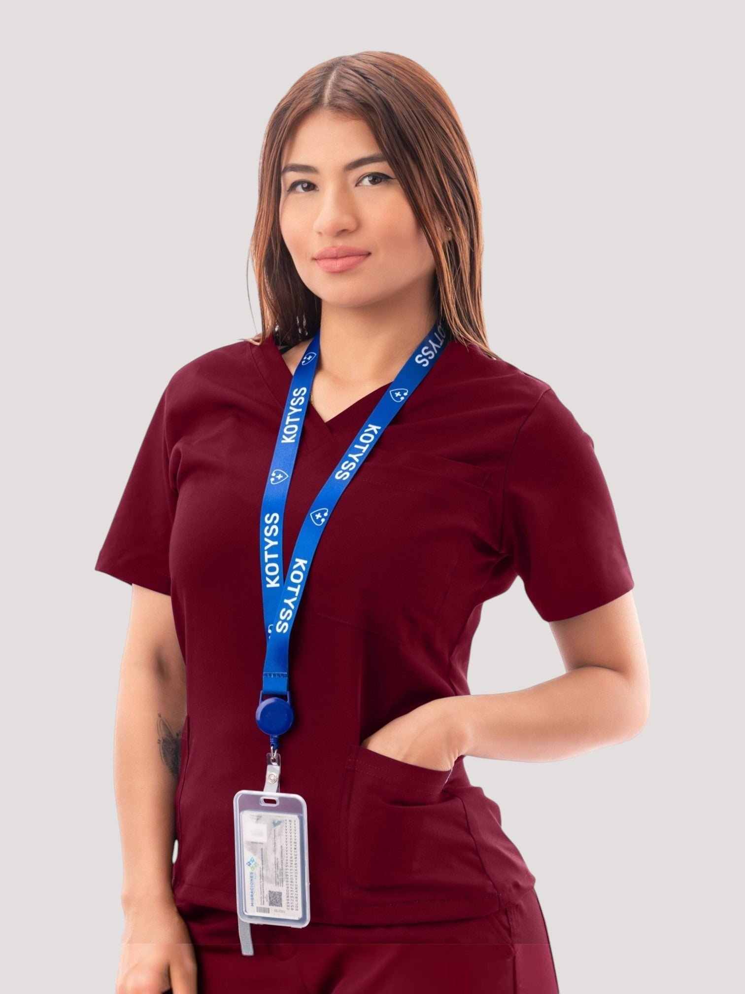 A woman wearing a burgundy Kotyss V-neck medical scrub top with pockets and a branded blue lanyard.